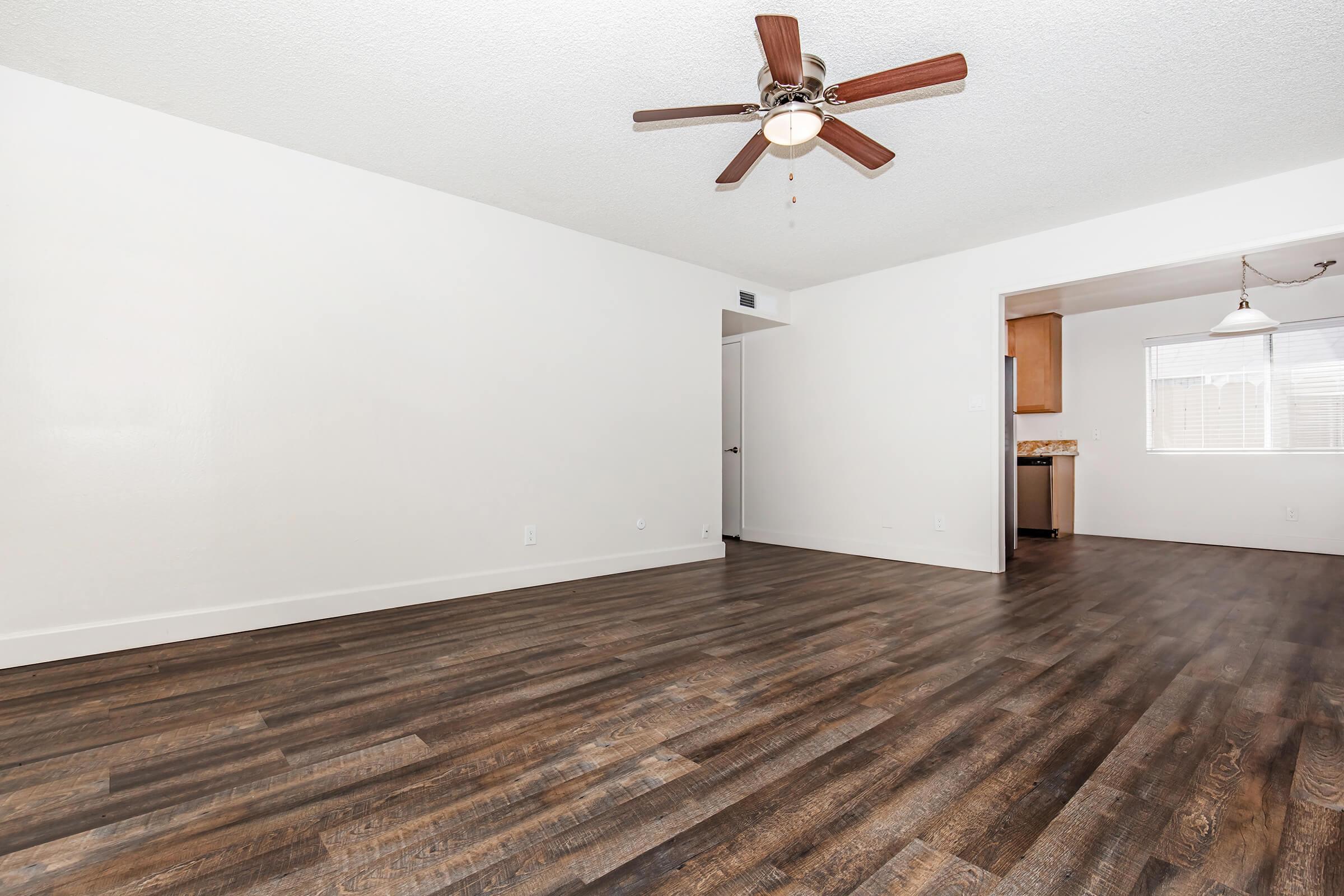 Spacious living area featuring hardwood-style flooring, a ceiling fan, and light-colored walls. On the right, a doorway leads to a kitchen area with natural lighting.