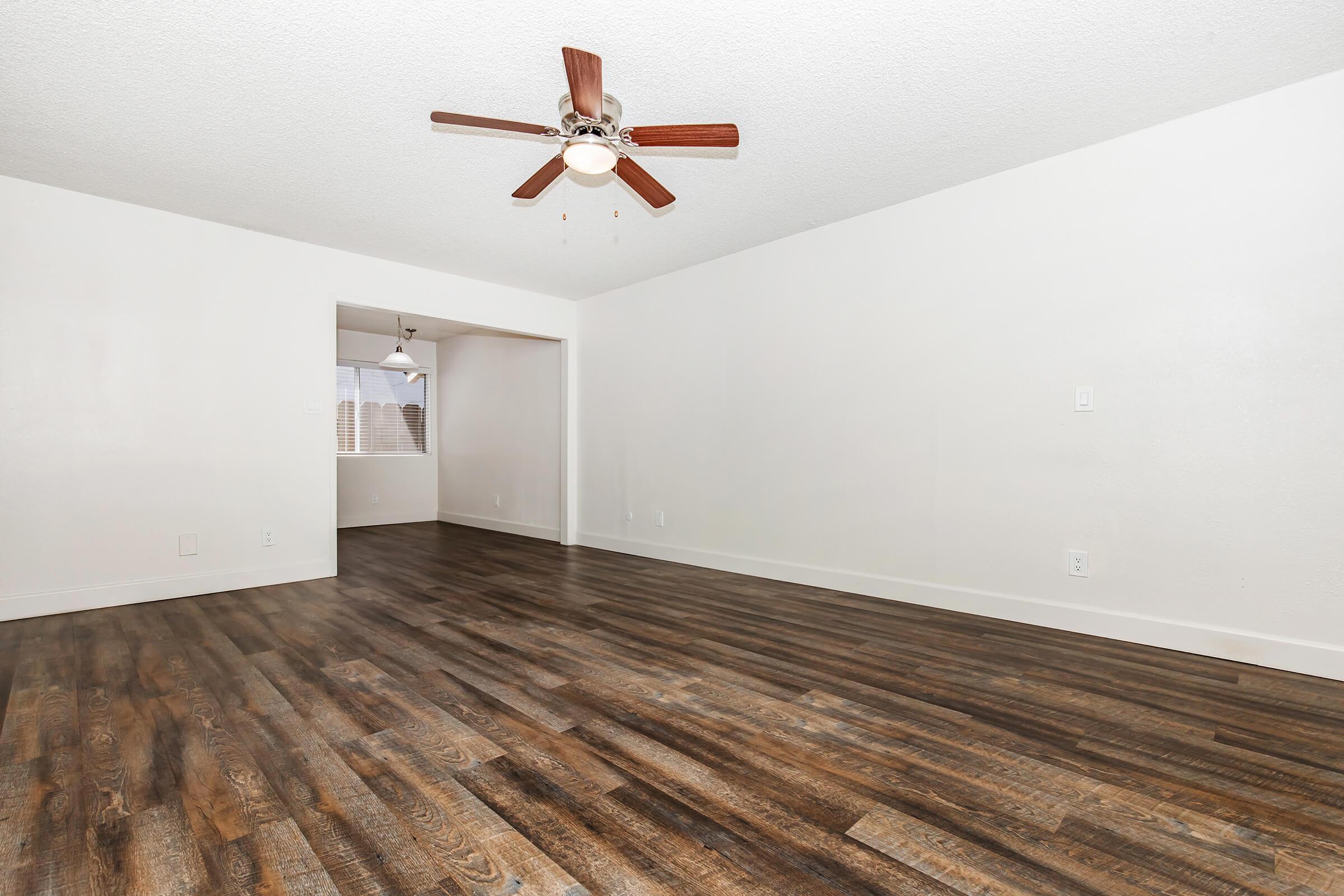 Spacious empty living room with light-colored walls and dark wood-style flooring. A ceiling fan with wooden blades hangs from the ceiling, and a doorway leads to another room in the background. Natural light comes from a window in the adjacent space.