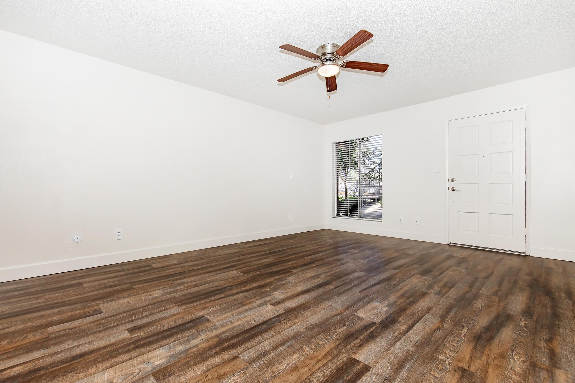A spacious, empty room with a ceiling fan, light brown laminate flooring, and white walls. A window with blinds lets in natural light, providing a view of the outdoors. The room features a simple door, enhancing the minimalist aesthetic.
