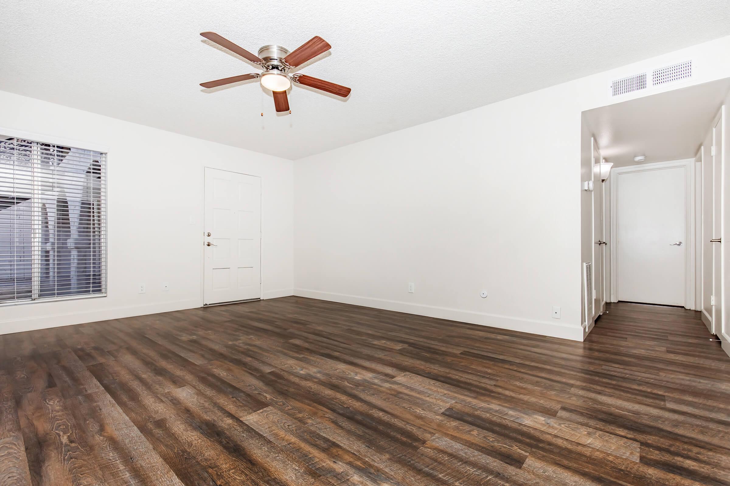 A spacious, empty room with a light-colored wall, wooden laminate flooring, and a ceiling fan. There's a window with blinds on the left side, a white door leading outside, and a hallway visible on the right. The overall ambiance is bright and open, ideal for various layouts and designs.