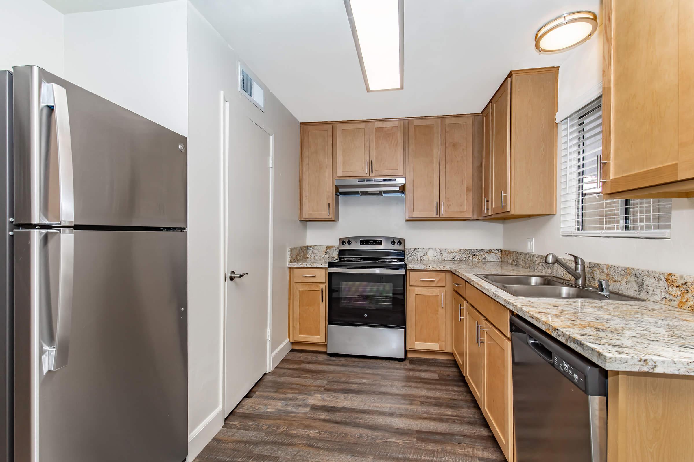 Modern kitchen featuring stainless steel appliances, including a refrigerator, oven, and dishwasher. Wooden cabinets offer storage space, complemented by a granite countertop. The room is well-lit with natural light from a window and overhead lighting, and has a sleek, contemporary design.