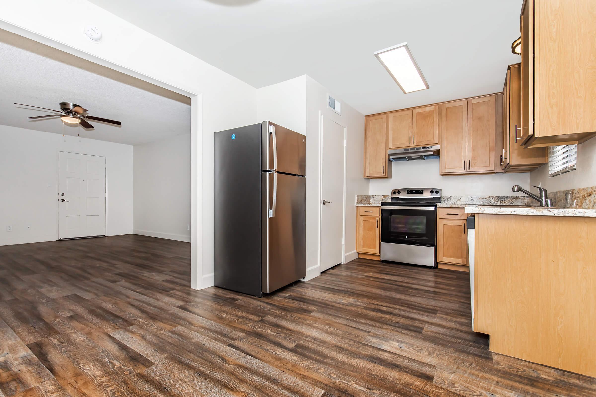 A modern kitchen area featuring a sleek black refrigerator and stainless steel appliances. The space has wooden flooring, light-colored cabinets, and a spacious layout, with a doorway leading to another room visible in the background.