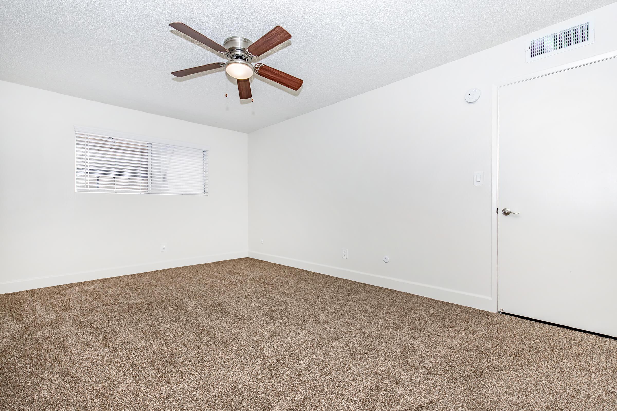 Spacious, empty room with light brown carpet and a white ceiling. A ceiling fan with wooden blades is installed. Natural light enters through a small window with blinds. There is a plain white door on one side, and the walls are painted a light color, creating a bright and neutral atmosphere.