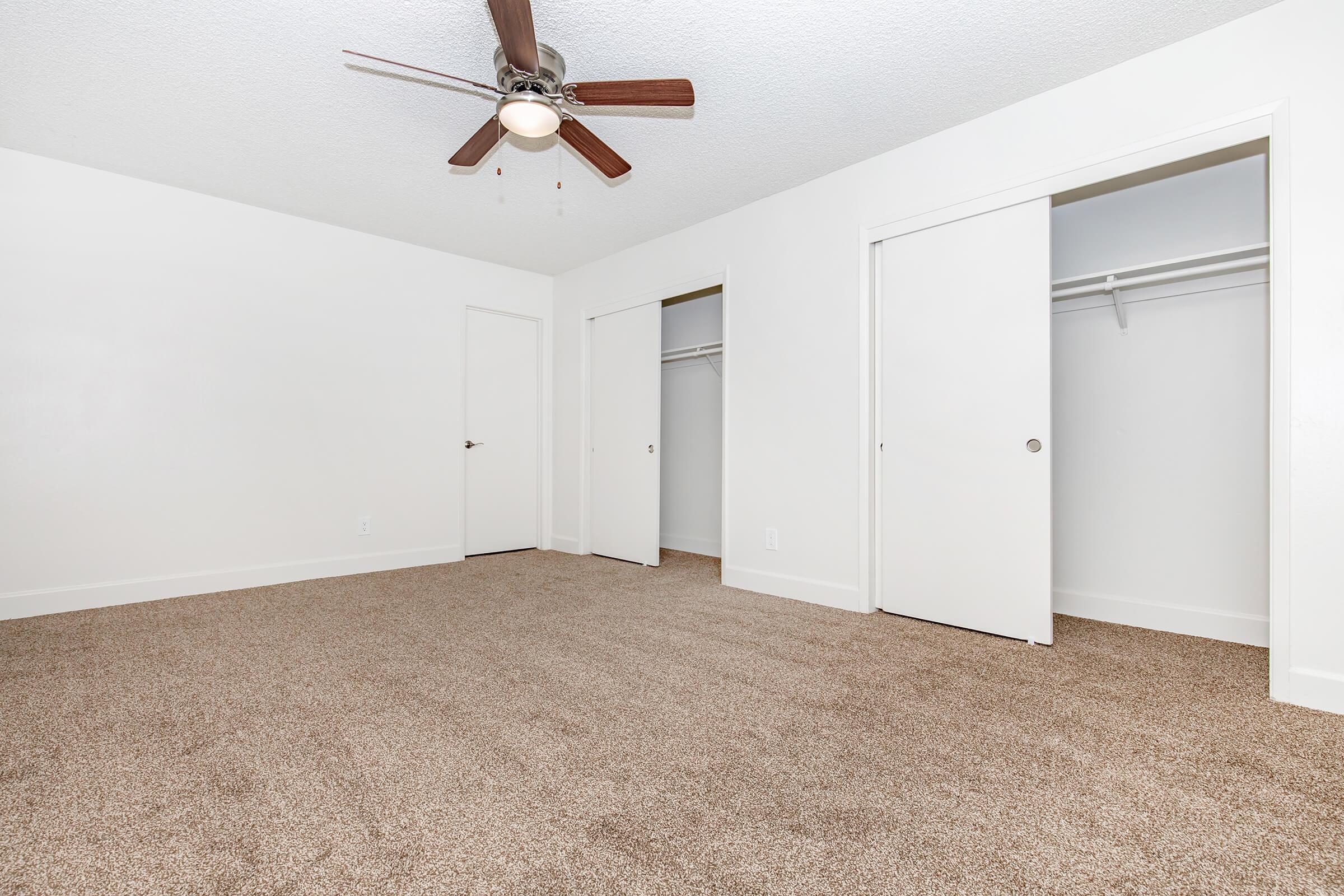 A spacious, empty bedroom featuring a light-colored carpet, white walls, and a ceiling fan. Two closet doors are visible on the right side, and a single door leads to another room or hallway. The room is bright and has an uncluttered appearance, ideal for personal customization.
