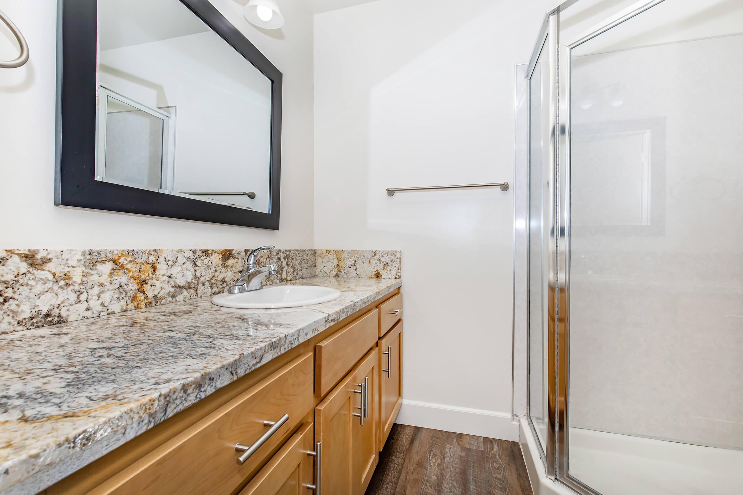A modern bathroom featuring a countertop with a sink and wooden cabinets. There is a large mirror above the sink and a glass shower enclosure with a white interior. The flooring is a dark wood finish, creating a contrast with the light-colored walls. Natural light illuminates the space.