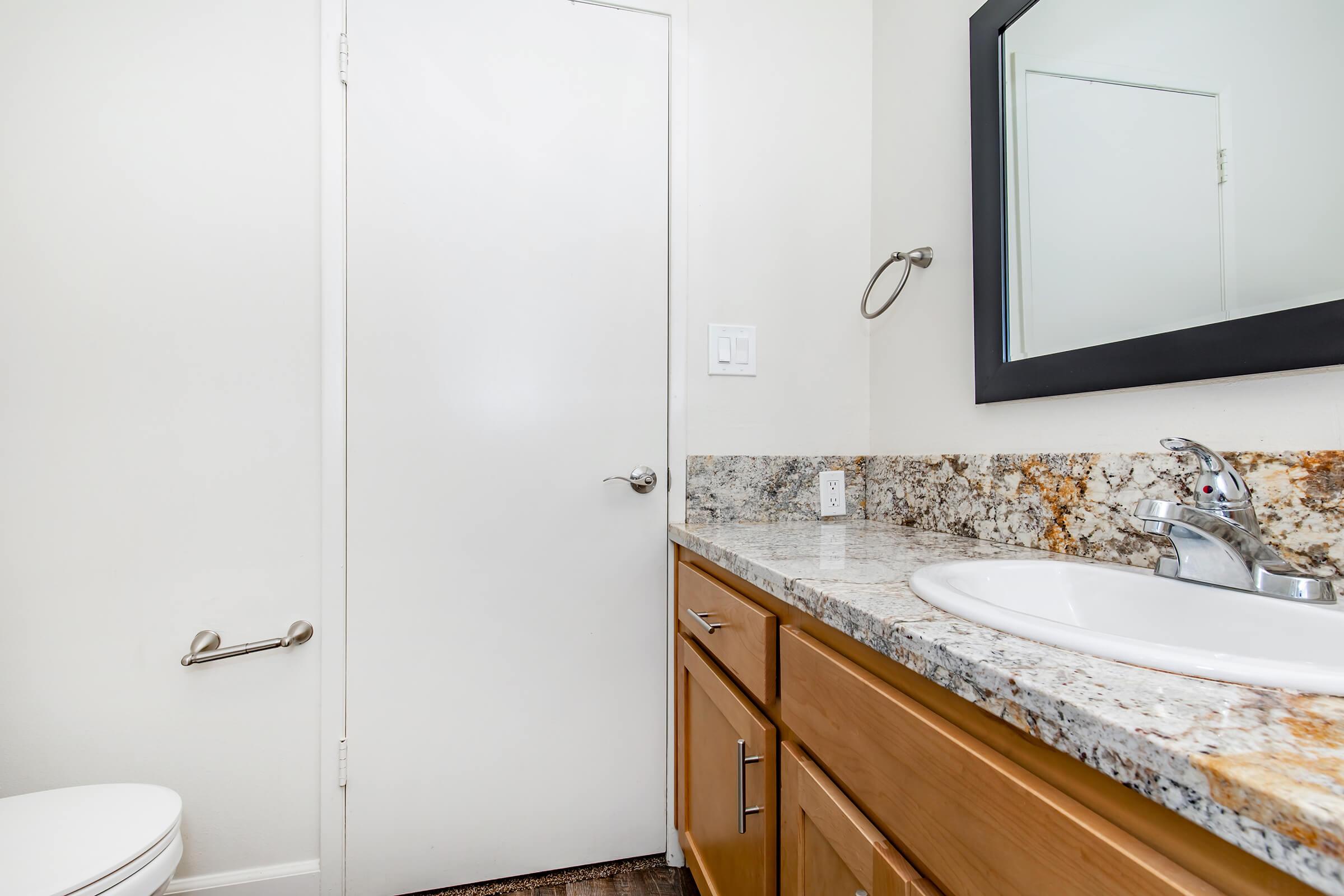 A modern bathroom featuring a white toilet, a granite countertop with a sink, wooden cabinets below, a mirror framed in black above the sink, and a door leading to another room. The walls are painted in a light color, and a towel bar is mounted next to the sink.