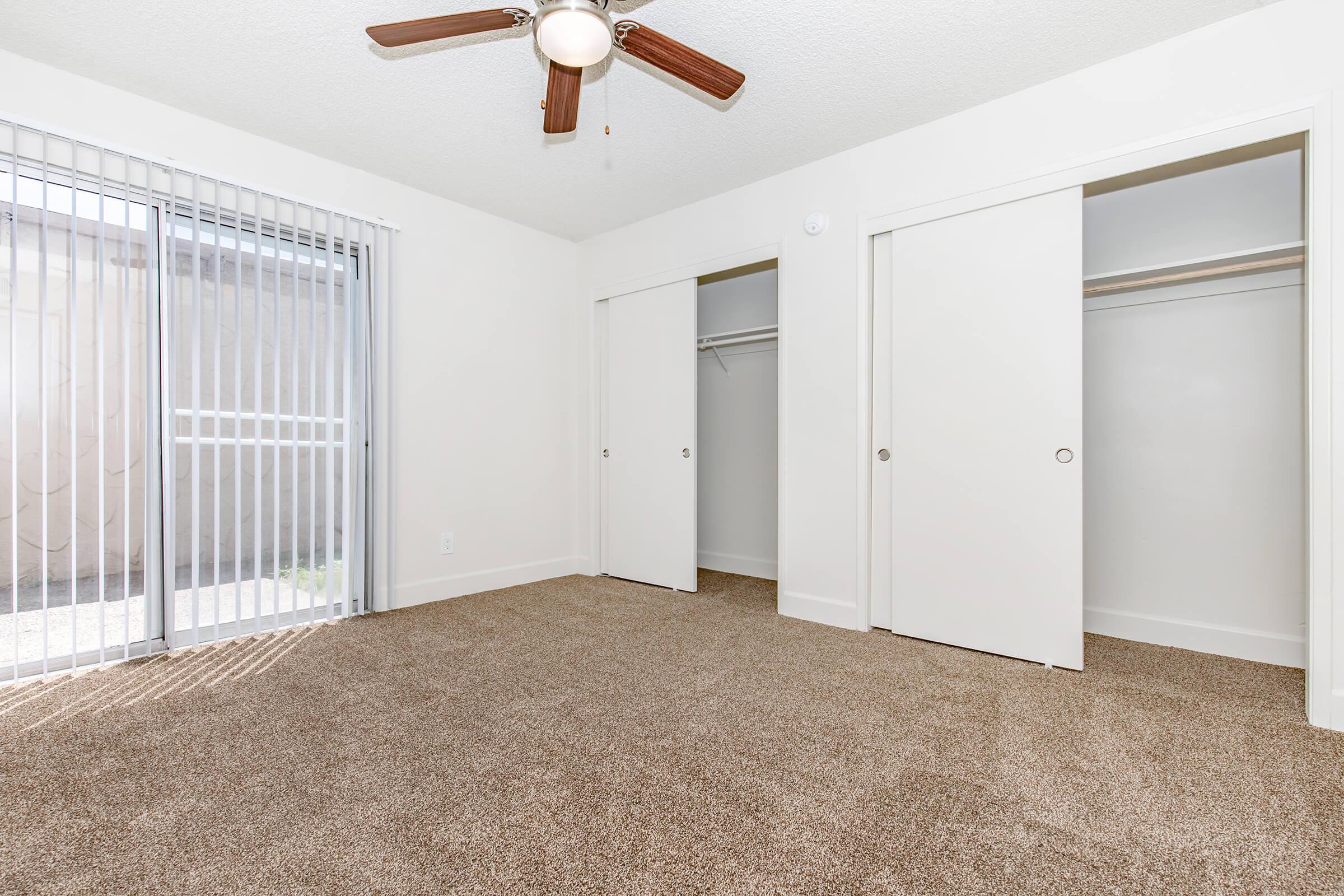 A spacious, well-lit bedroom featuring beige carpet flooring, a ceiling fan, and two white sliding closet doors. There are vertical blinds on a large window letting in natural light, with an outdoor view visible. The walls are painted in a light color, enhancing the brightness of the room.