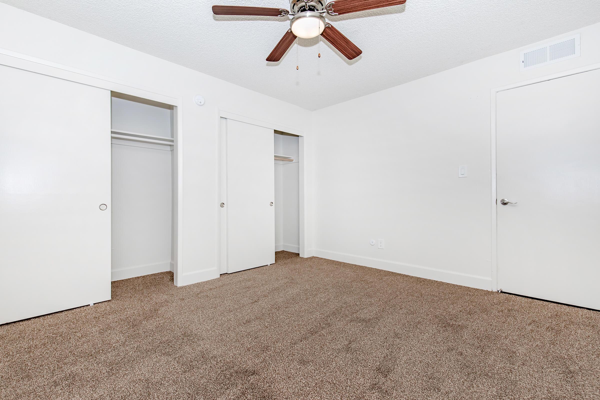 A well-lit room featuring beige carpet flooring and white walls. The space includes three closed doors leading to closets, a ceiling fan with wooden blades, and a light fixture. The room is empty and appears ready for furnishing.