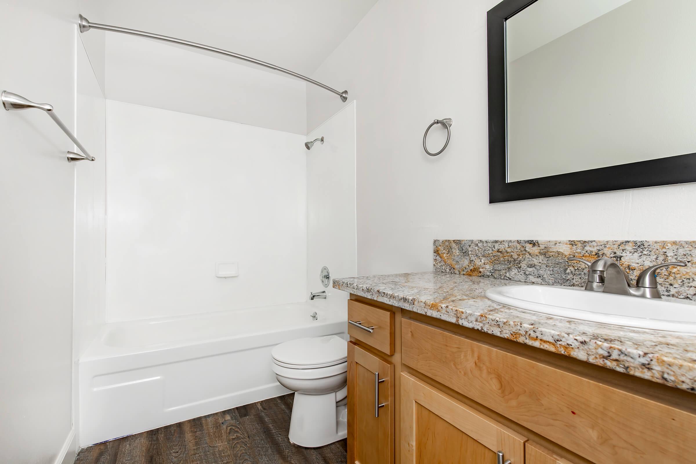 A clean bathroom featuring a shower/tub combination, a modern sink with a wooden cabinet, a round mirror, and a towel rack. The walls are painted white, and the floor has dark wood laminate.