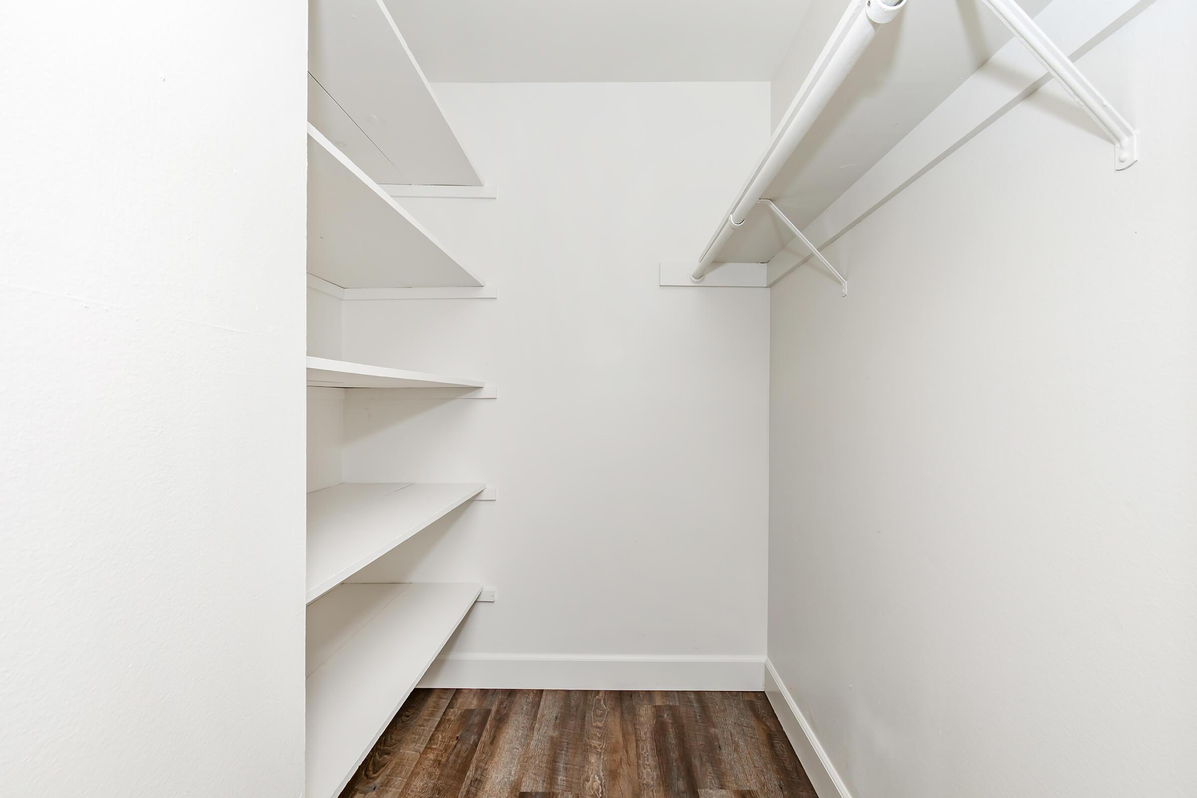 Empty closet with white walls and shelves on one side, featuring a wooden floor. The opposite side has a hanging rod for clothes, creating a spacious, minimalist storage area.