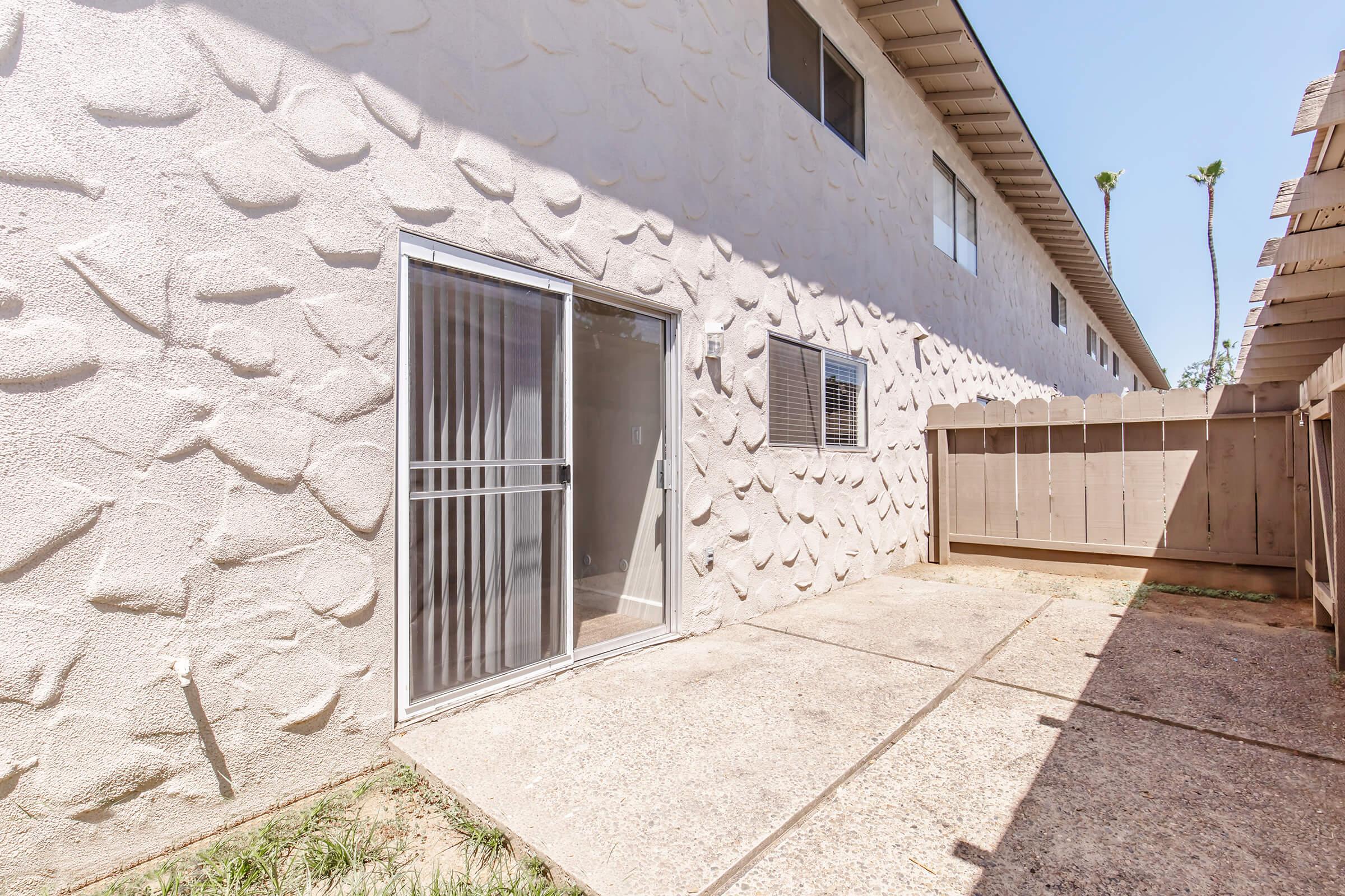 A view of a residential backyard with a concrete patio area. The wall features a textured, light-colored surface, and there are sliding glass doors leading to an interior space. A wooden fence borders one side of the area, and palm trees are visible in the background under a clear blue sky.