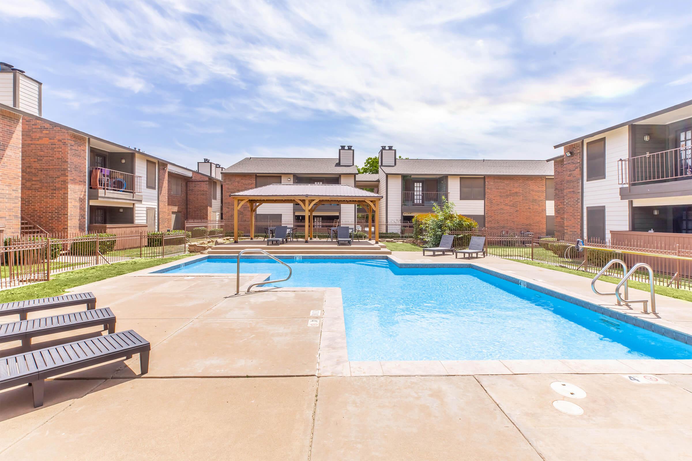 A well-maintained outdoor swimming pool surrounded by lounge chairs and a shaded pergola. The pool area is bordered by landscaped green spaces and two-story apartment buildings with brick facades in the background. The sky is clear with a few clouds, creating a bright and inviting atmosphere.