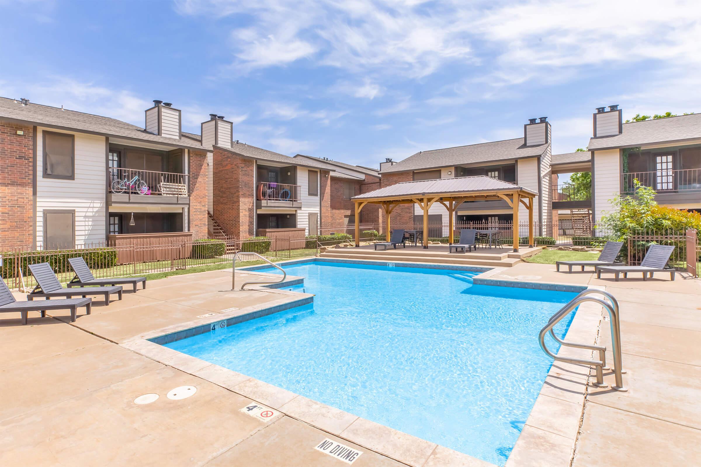 A sunny outdoor swimming pool surrounded by lounge chairs and a wooden gazebo, with apartment buildings in the background. Clear blue sky and well-maintained landscaping create a relaxing atmosphere.