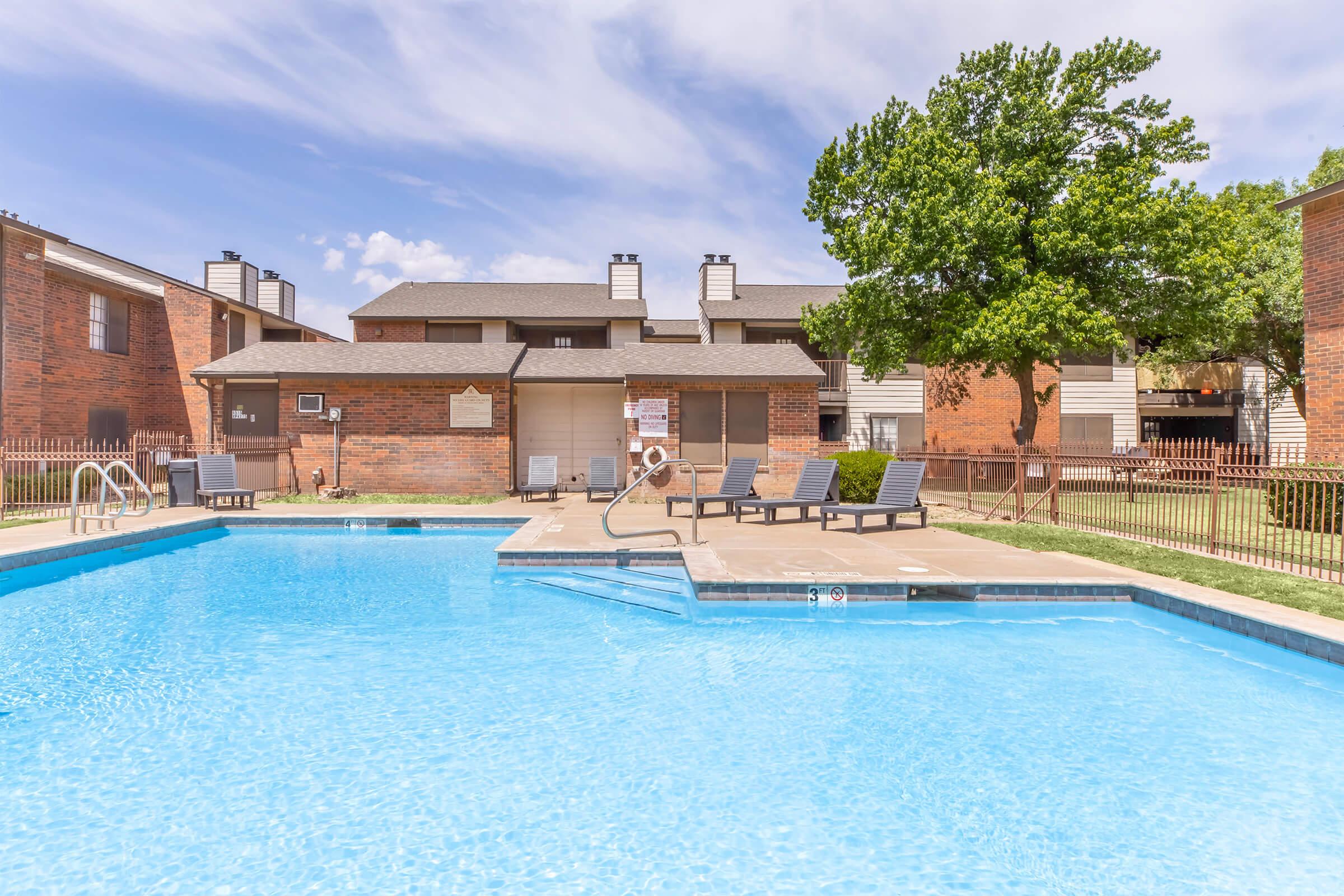 A bright outdoor swimming pool surrounded by lounge chairs and a green lawn, with a brick building in the background. The sky is clear with scattered clouds, creating a relaxing atmosphere for leisure and recreation.