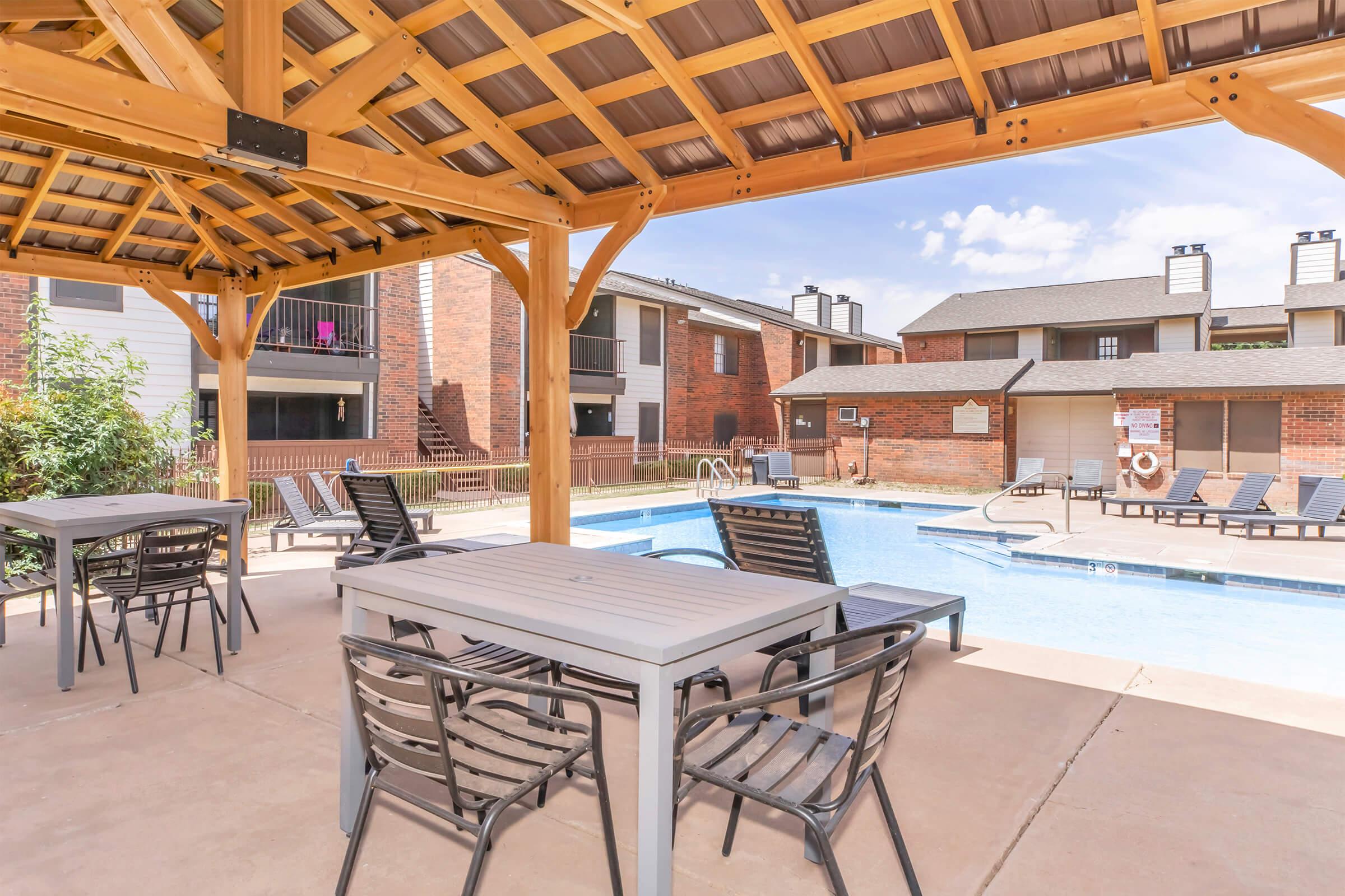 A shaded outdoor area with tables and chairs near a swimming pool. The pool is surrounded by lounge chairs and features a backdrop of residential buildings. The sky is clear with a few clouds, creating a bright and inviting atmosphere.
