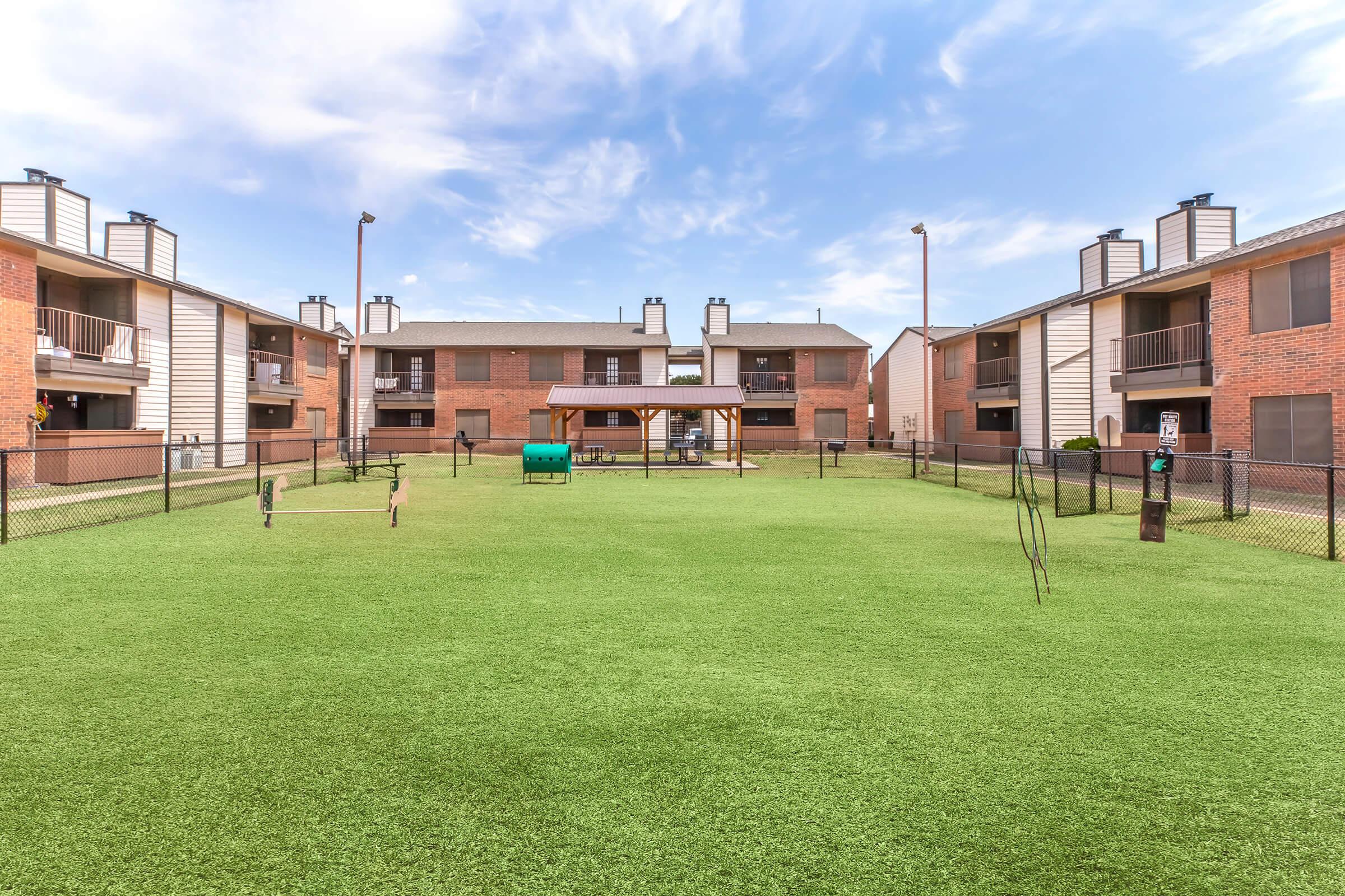 A spacious green courtyard surrounded by apartment buildings. The area features a small playground with swings and a picnic shelter, enclosed by a black fence. The sky is clear with a few clouds, providing a bright and inviting atmosphere.