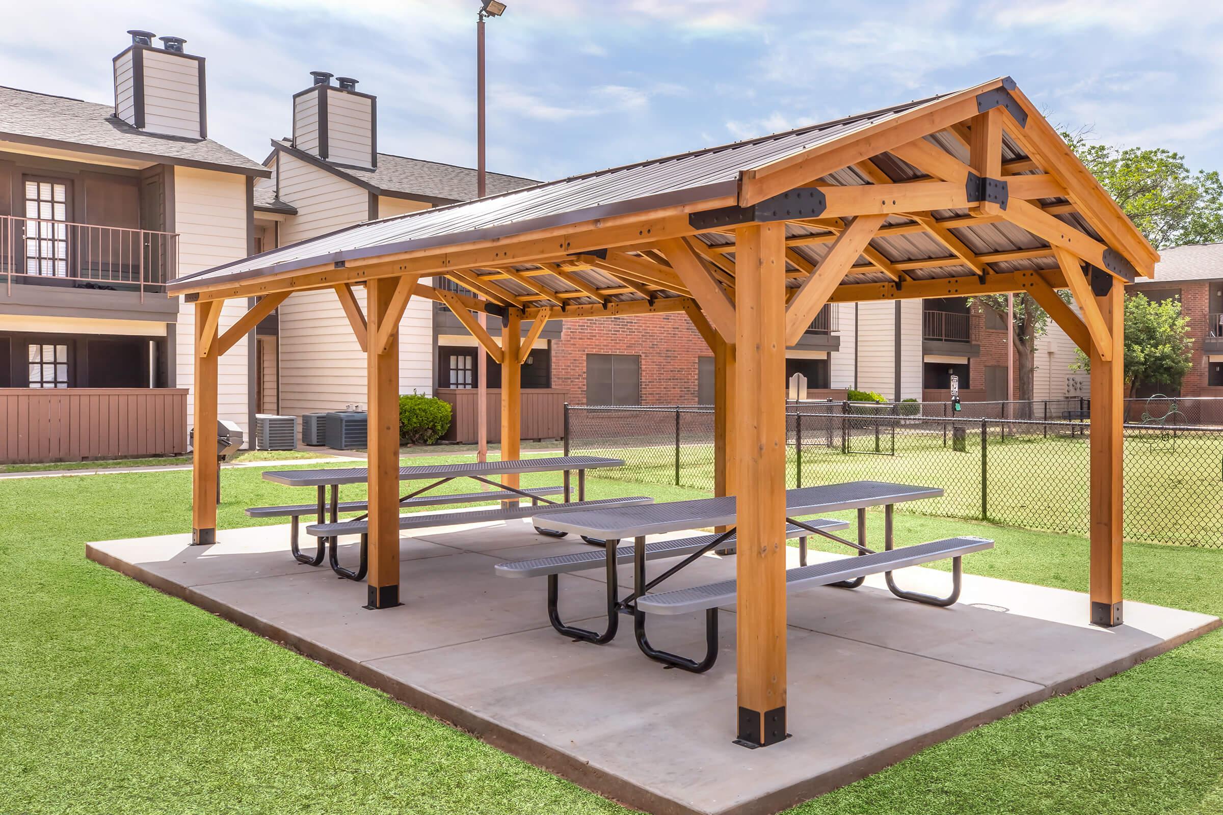 A wooden pavilion with a sloped roof provides shade over several picnic tables, set on a concrete surface. The structure is located in a grassy area, surrounded by an apartment complex and a fence. The scene is bright and inviting, ideal for outdoor gatherings.