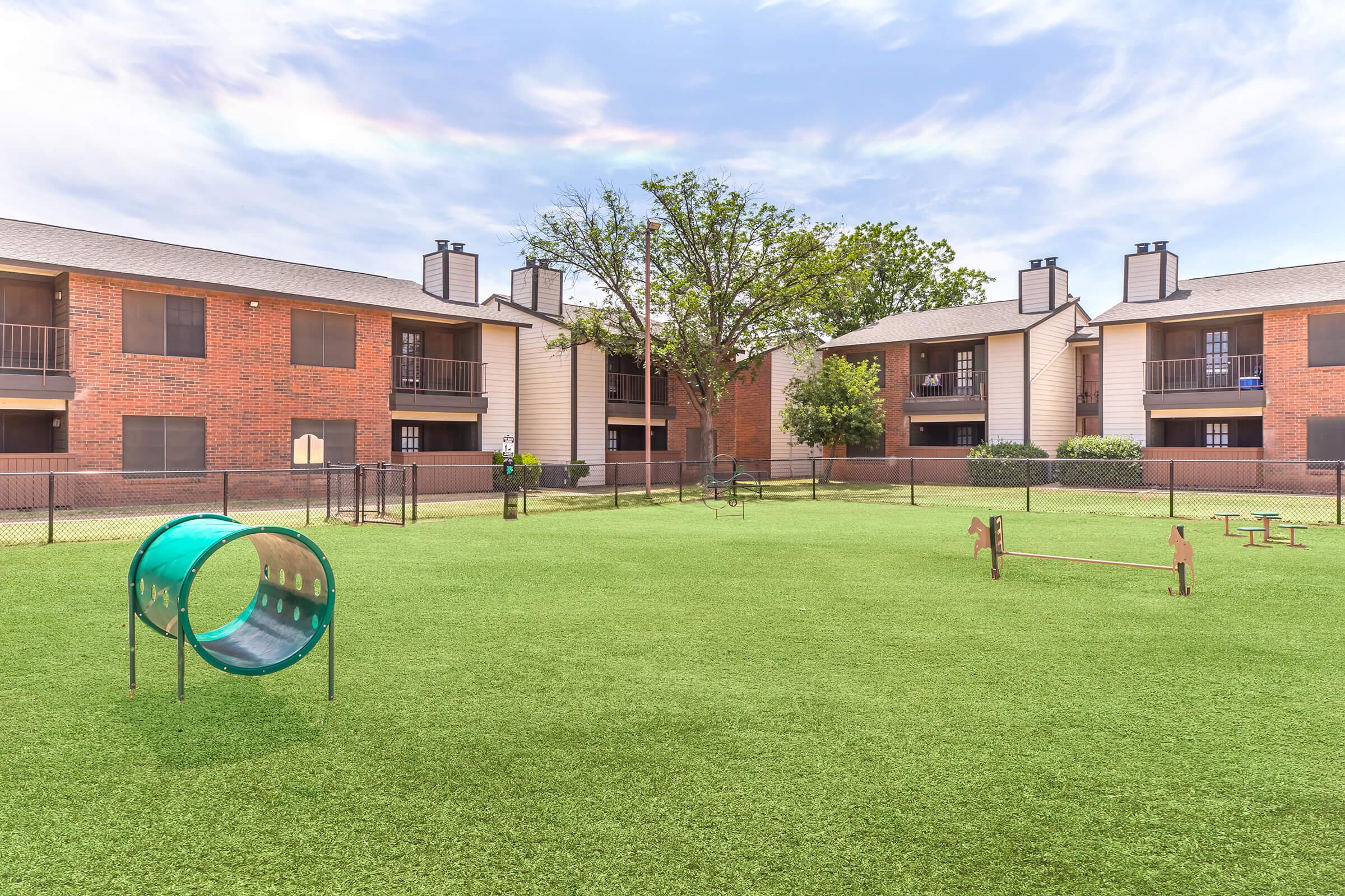 A grassy dog park area surrounded by brick apartment buildings. The park features agility equipment, including a tunnel and small jumps, along with a few trees and a fenced perimeter. The sky is bright with scattered clouds, creating a welcoming outdoor space for pets.