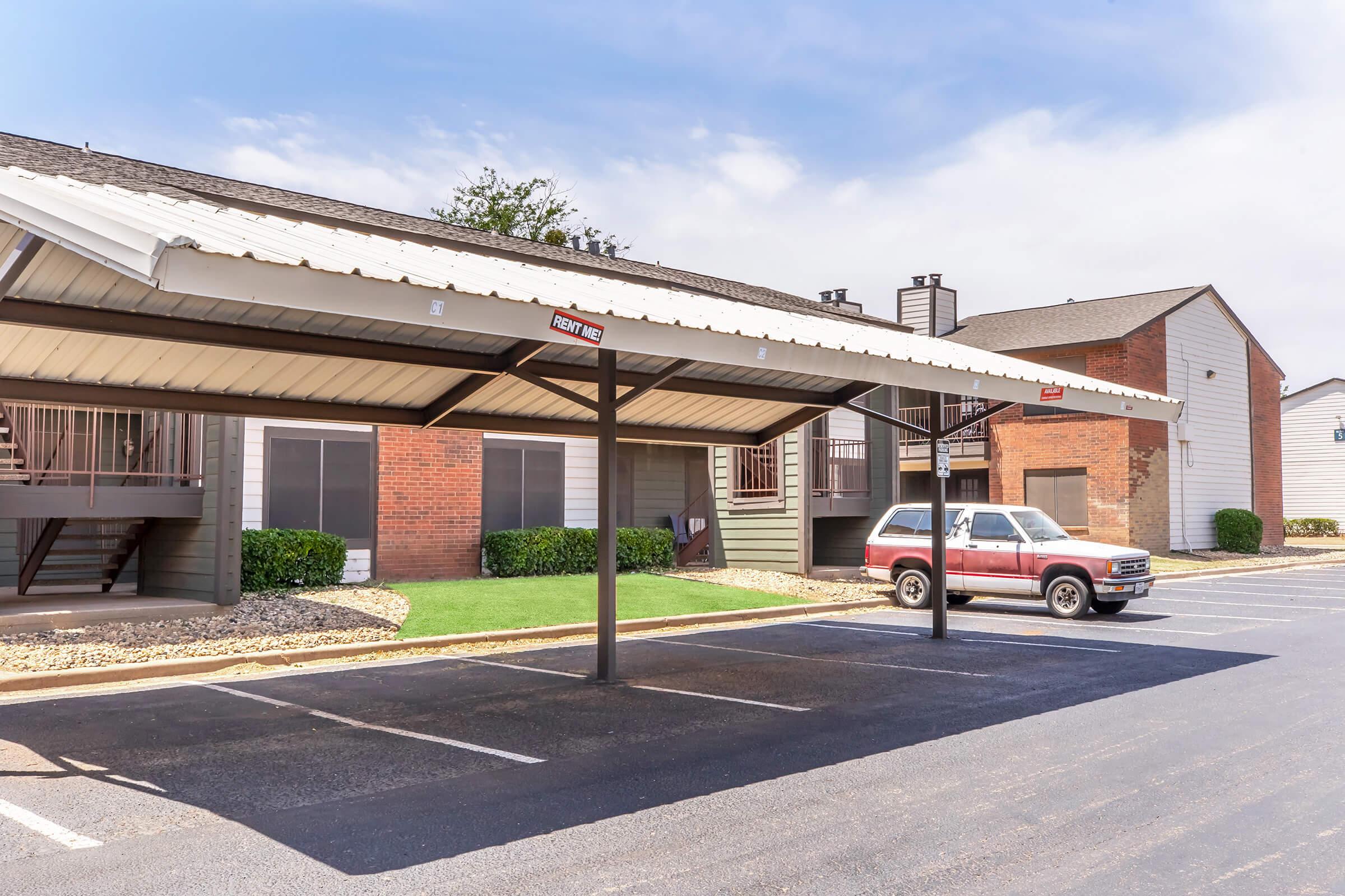A parking area featuring covered carports next to an apartment complex. There are several empty parking spaces, a red truck parked under one of the carports, and a neatly maintained lawn with shrubs. The sky is partly cloudy, and the buildings have a mix of brick and siding exteriors.