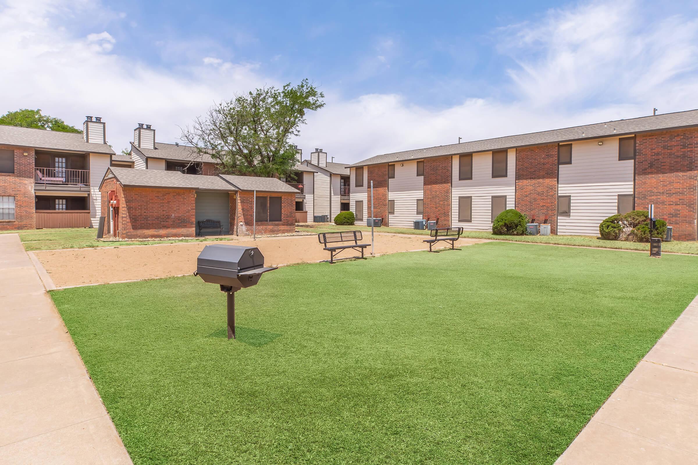 A landscaped courtyard in an apartment complex featuring green artificial grass, picnic tables, and a charcoal grill. Surrounding buildings are two stories tall, with a mix of red brick and white siding. The sky is partly cloudy, creating a bright and inviting atmosphere.