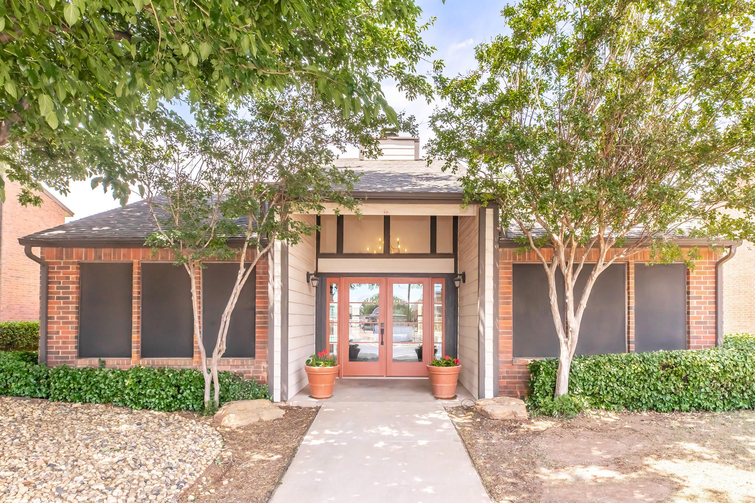 A charming single-story building with a front entrance featuring large windows and a double door. It is framed by leafy trees and shrubs, with flower pots on either side of the entrance. The exterior combines brick and siding, set in a well-maintained landscape.