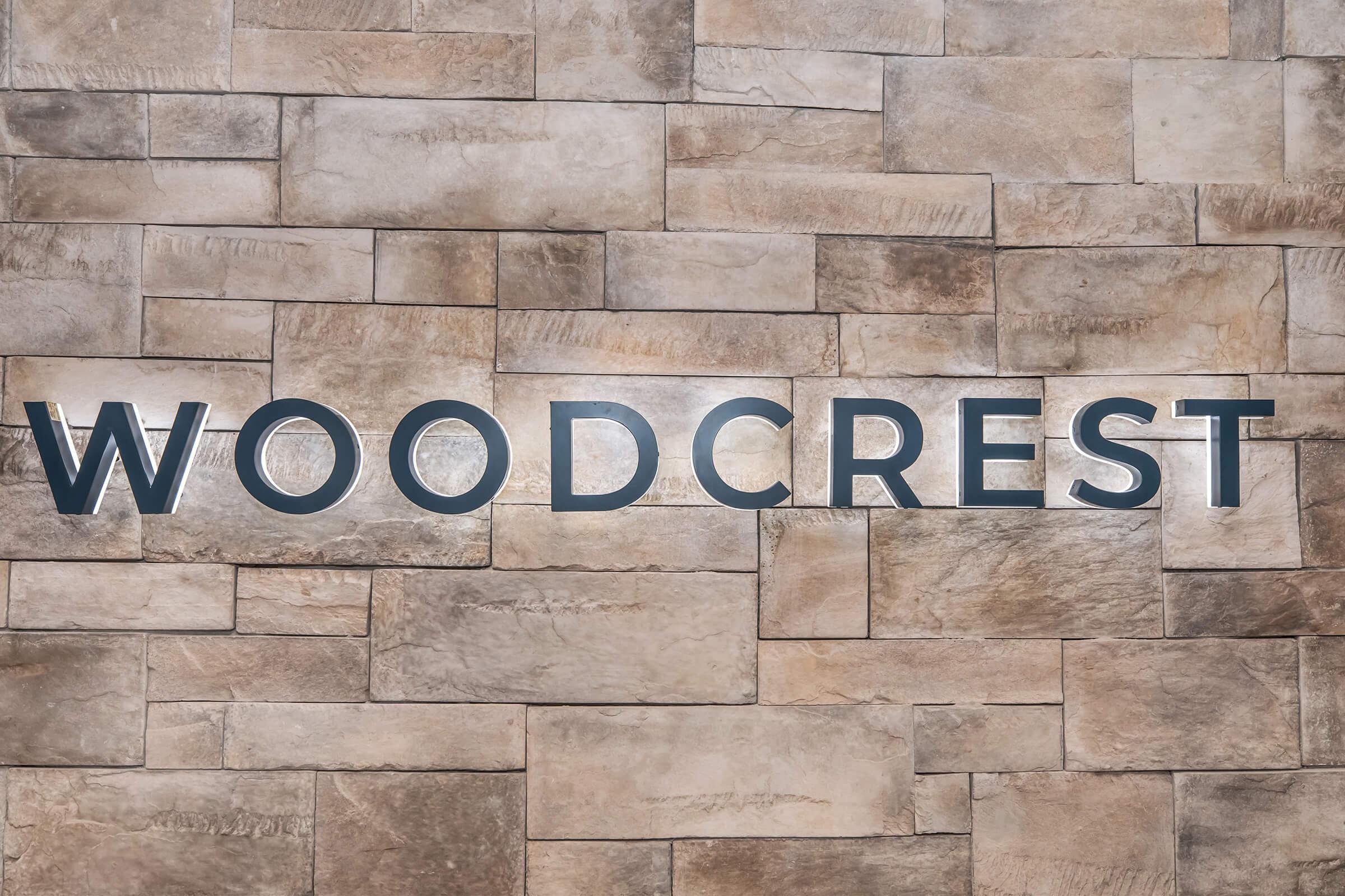 Sign displaying the word "WOODCREST" in bold letters, set against a textured stone wall with a mix of light and warm tones. The letters are dark and slightly raised, creating a contrast with the natural stone background.