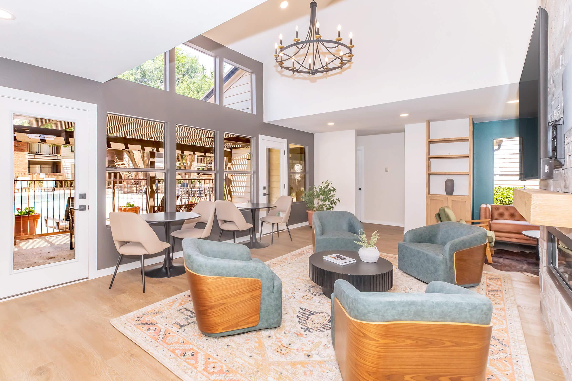 A modern lounge area featuring four teal armchairs with wooden accents, surrounded by large windows. A round black coffee table sits in the center on a patterned rug. A chandelier hangs from the ceiling, and there's a cozy corner with a brown chair and shelves. Natural light fills the space, enhancing the inviting atmosphere.