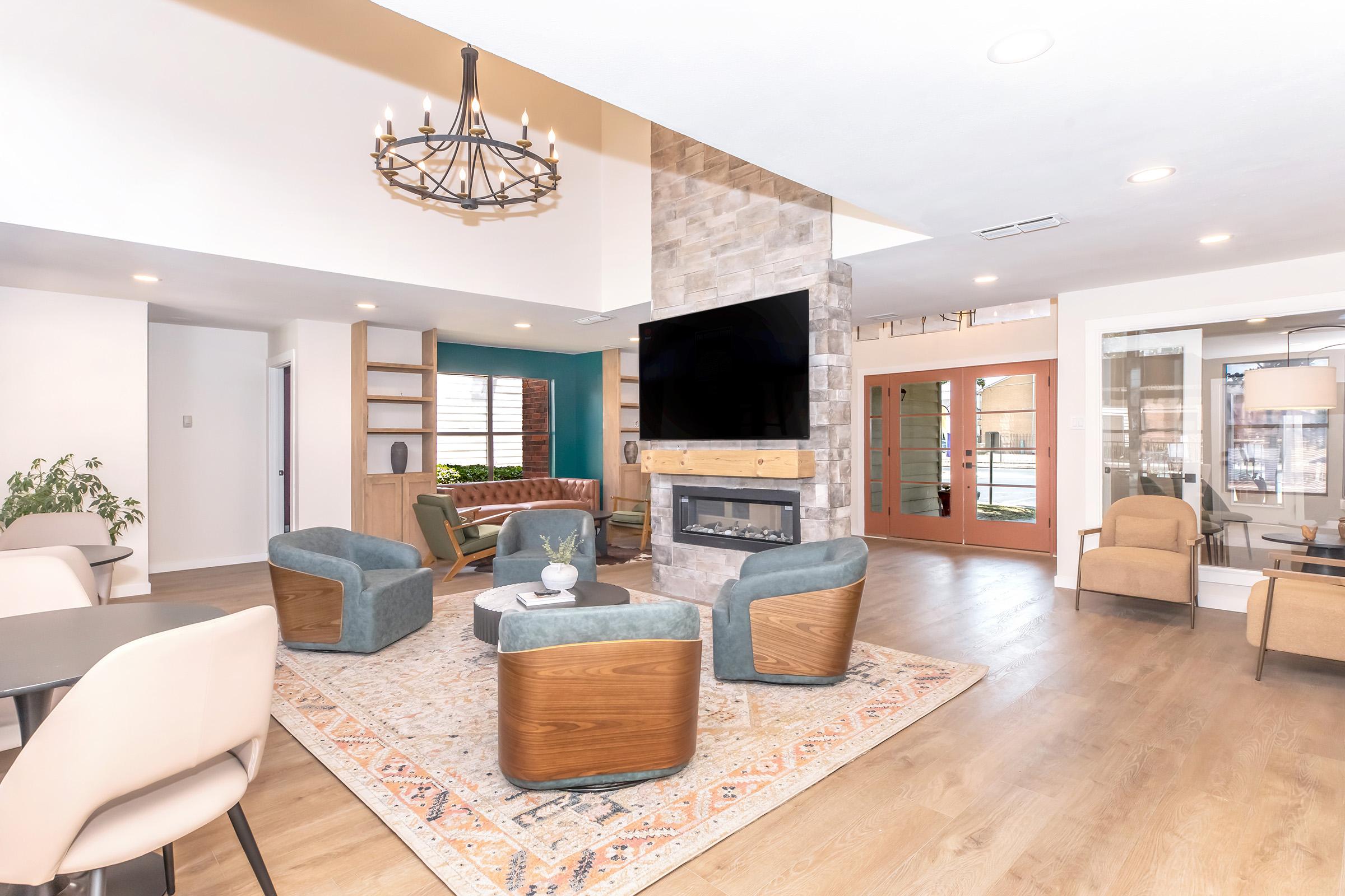 A modern living space featuring a cozy seating area with blue and brown armchairs around a central coffee table. A stone fireplace with a television above it serves as a focal point, complemented by a stylish chandelier. The room has large windows and a warm, inviting atmosphere with a decorative area rug.