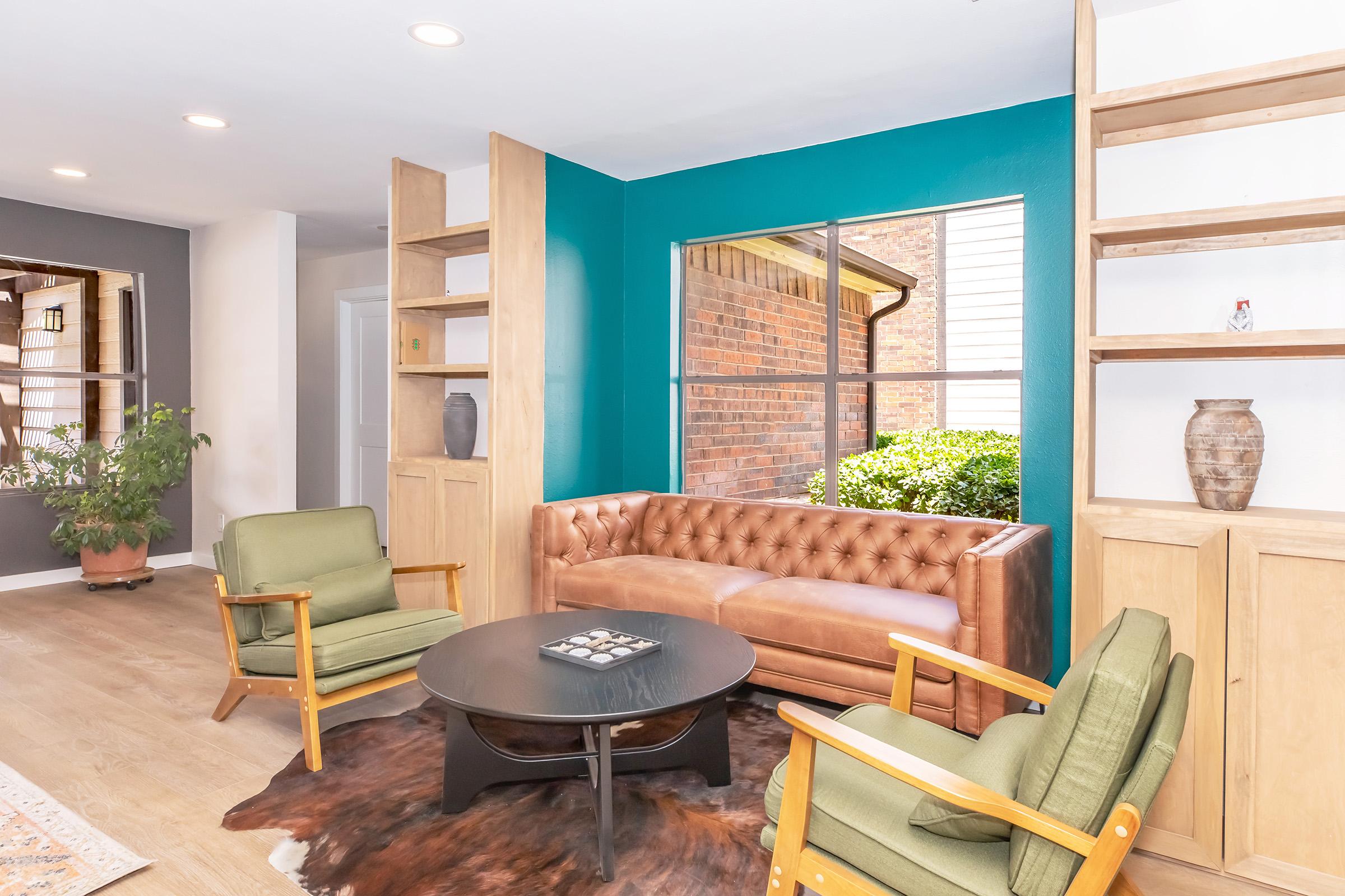 A cozy living room featuring a brown tufted leather sofa, two green armchairs, and a round black coffee table. The room has wooden shelves with decorative items and a large window with a view of greenery outside. The walls are painted a vibrant teal, adding a pop of color to the space.