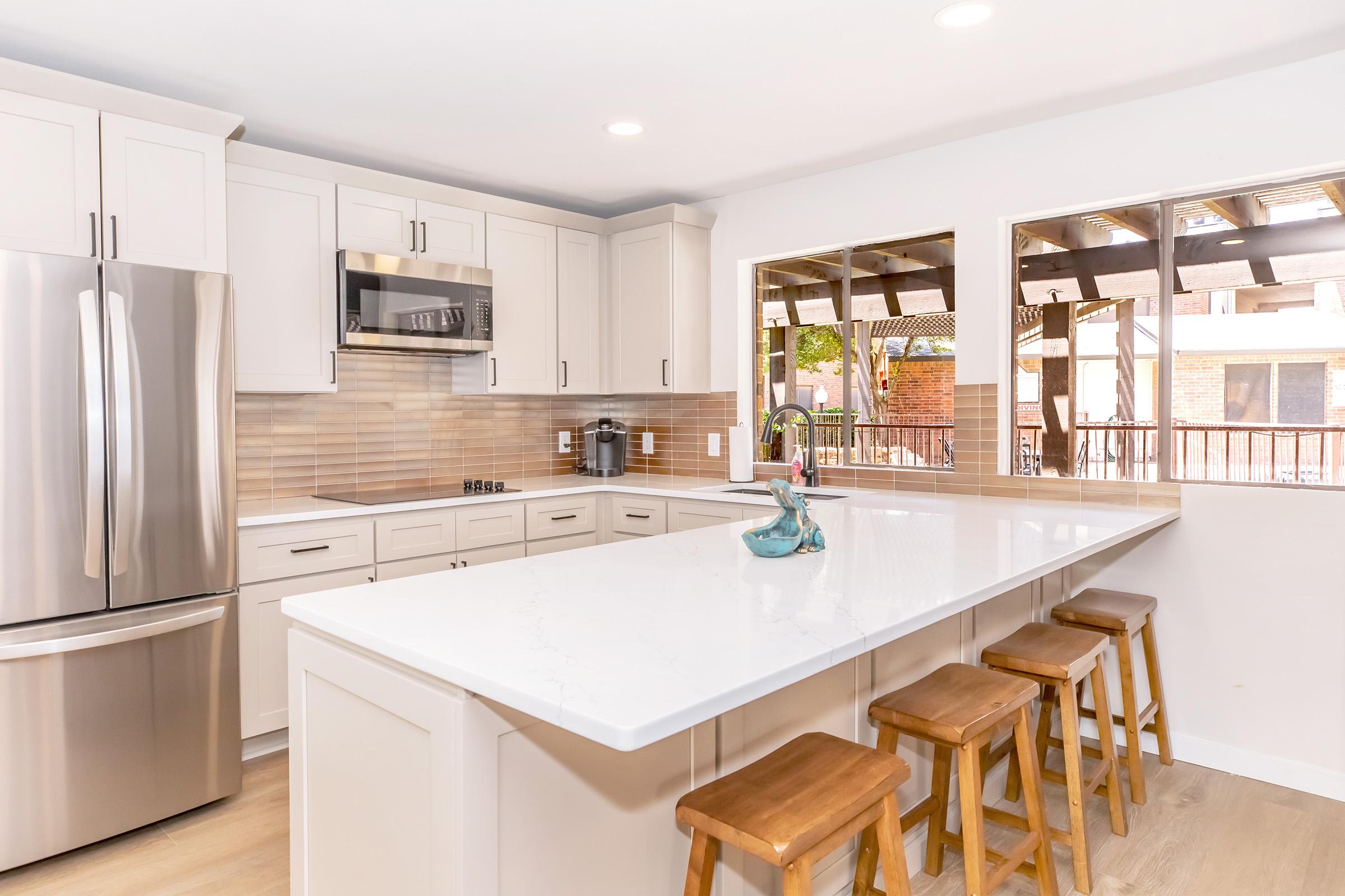 Modern kitchen featuring white cabinetry, stainless steel appliances, and a marble island with wooden stools. Large windows allow natural light, framing a view of an outdoor patio area. The backsplash has warm, neutral tones that complement the overall design.