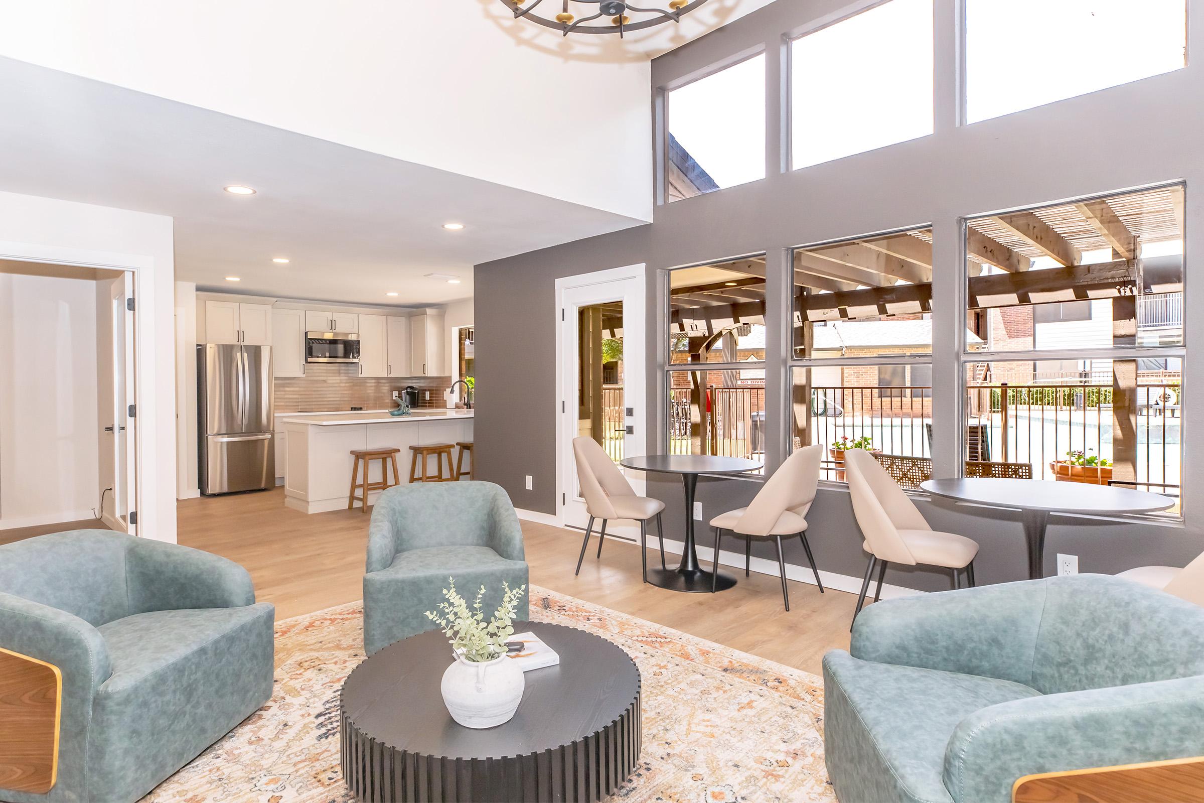 A modern living space featuring a cozy seating area with green chairs and a round coffee table. Large windows allow natural light, showcasing a contemporary kitchen in the background. The decor includes plants and a stylish area rug, creating a welcoming and inviting atmosphere.