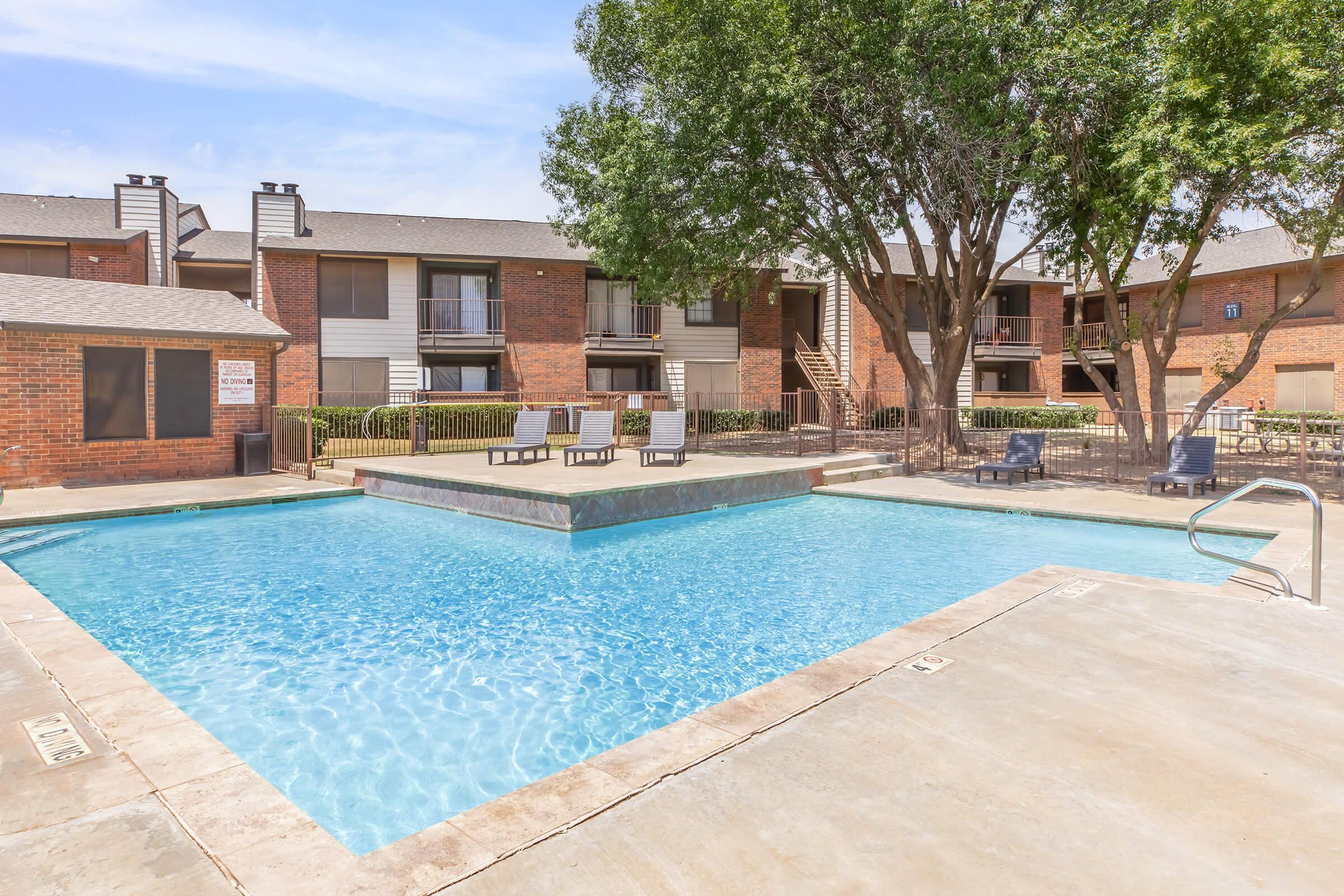 Swimming pool surrounded by lounge chairs and apartment buildings. Trees provide shade in a relaxing outdoor setting. The pool features a clear blue water and a stone deck, creating an inviting atmosphere for leisure and enjoyment.