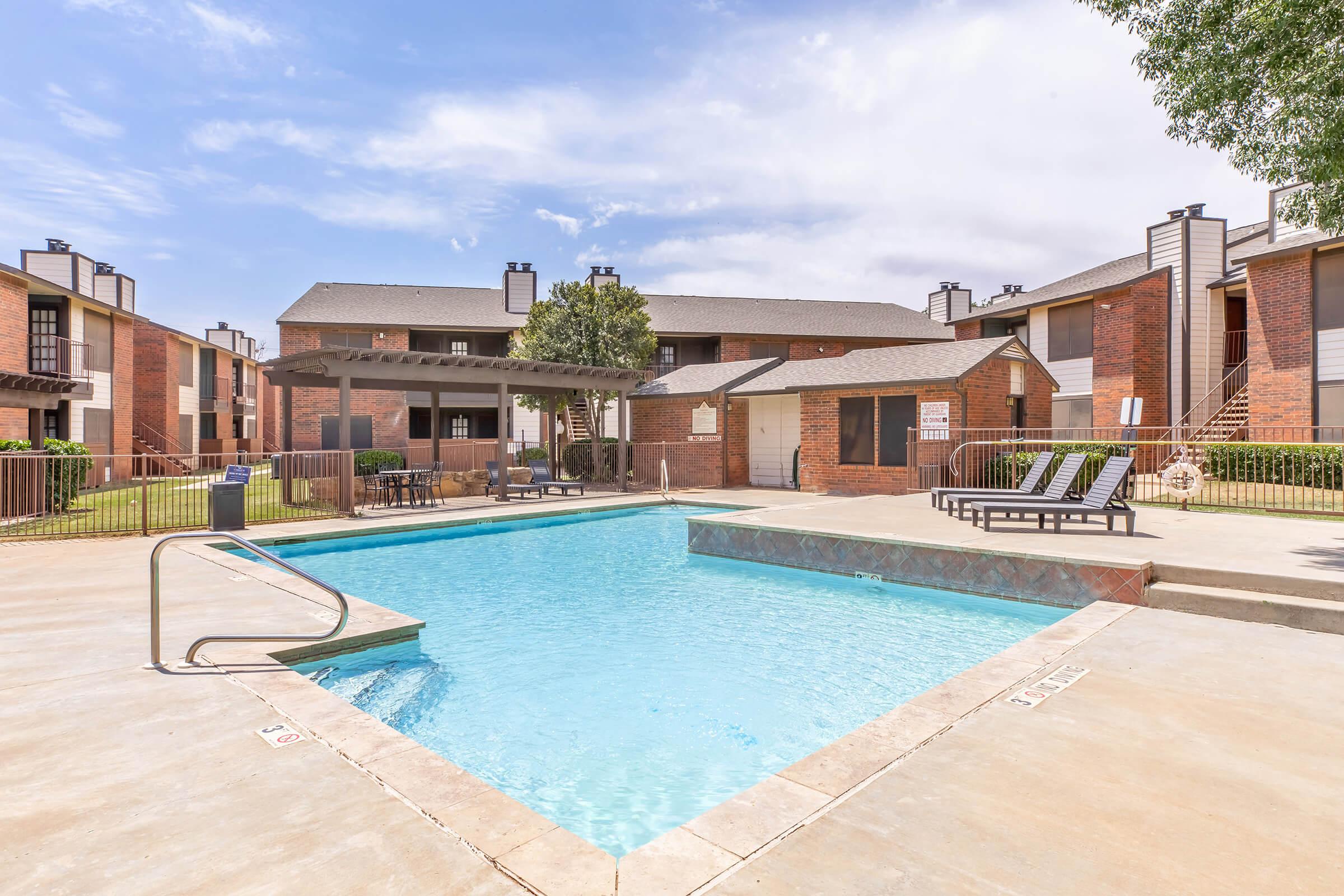 A view of a landscaped outdoor pool area surrounded by apartment buildings. The pool features a diving board and is bordered by lounge chairs. There is a shaded gazebo nearby and well-maintained green grass, with trees creating a pleasant atmosphere under a clear blue sky.