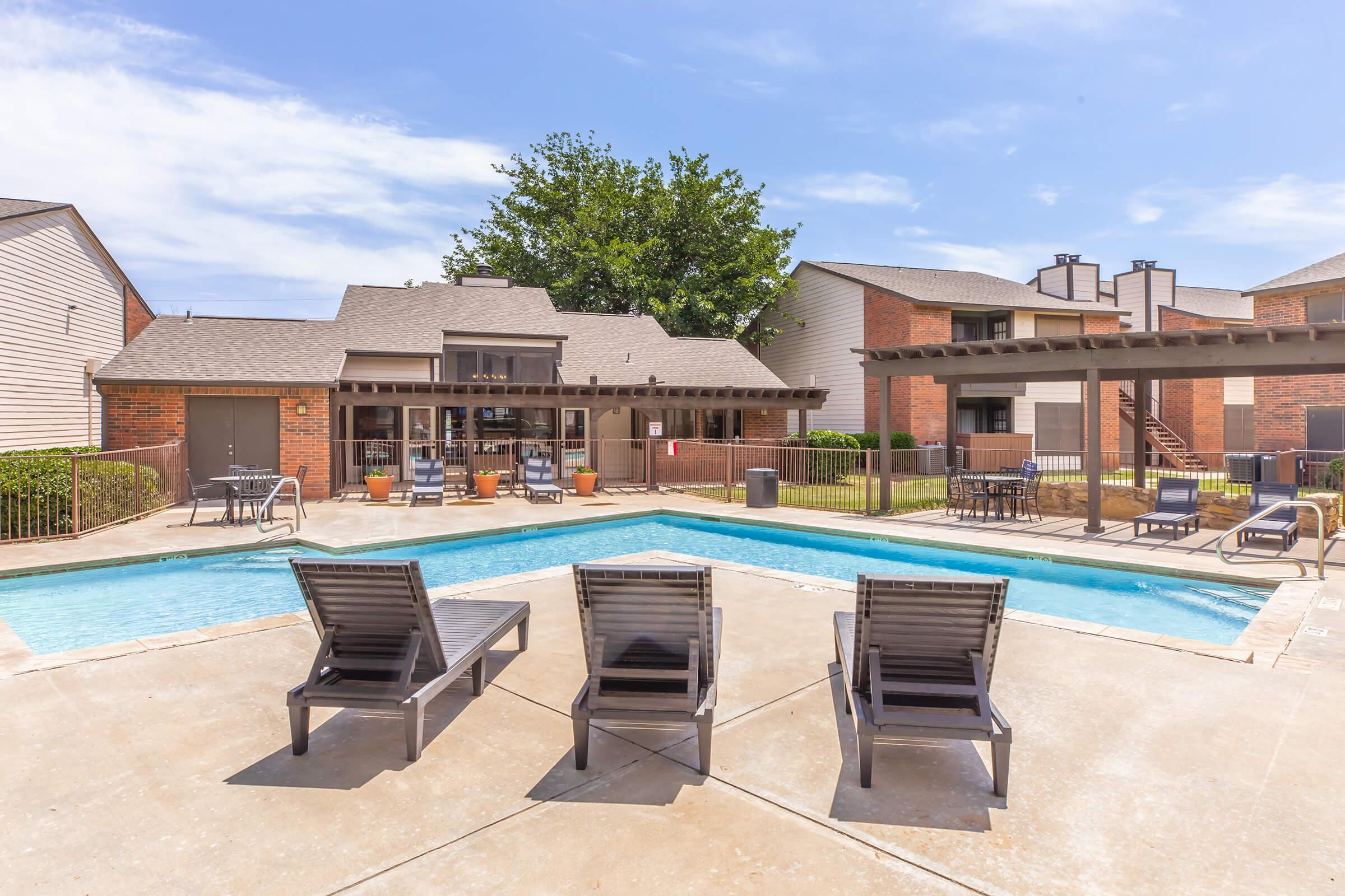 A tranquil swimming pool area surrounded by lounge chairs, with a well-maintained patio and green landscaping. Nearby, a building features large windows and a covered seating area. The sky is blue with a few clouds, creating a peaceful atmosphere for relaxation.