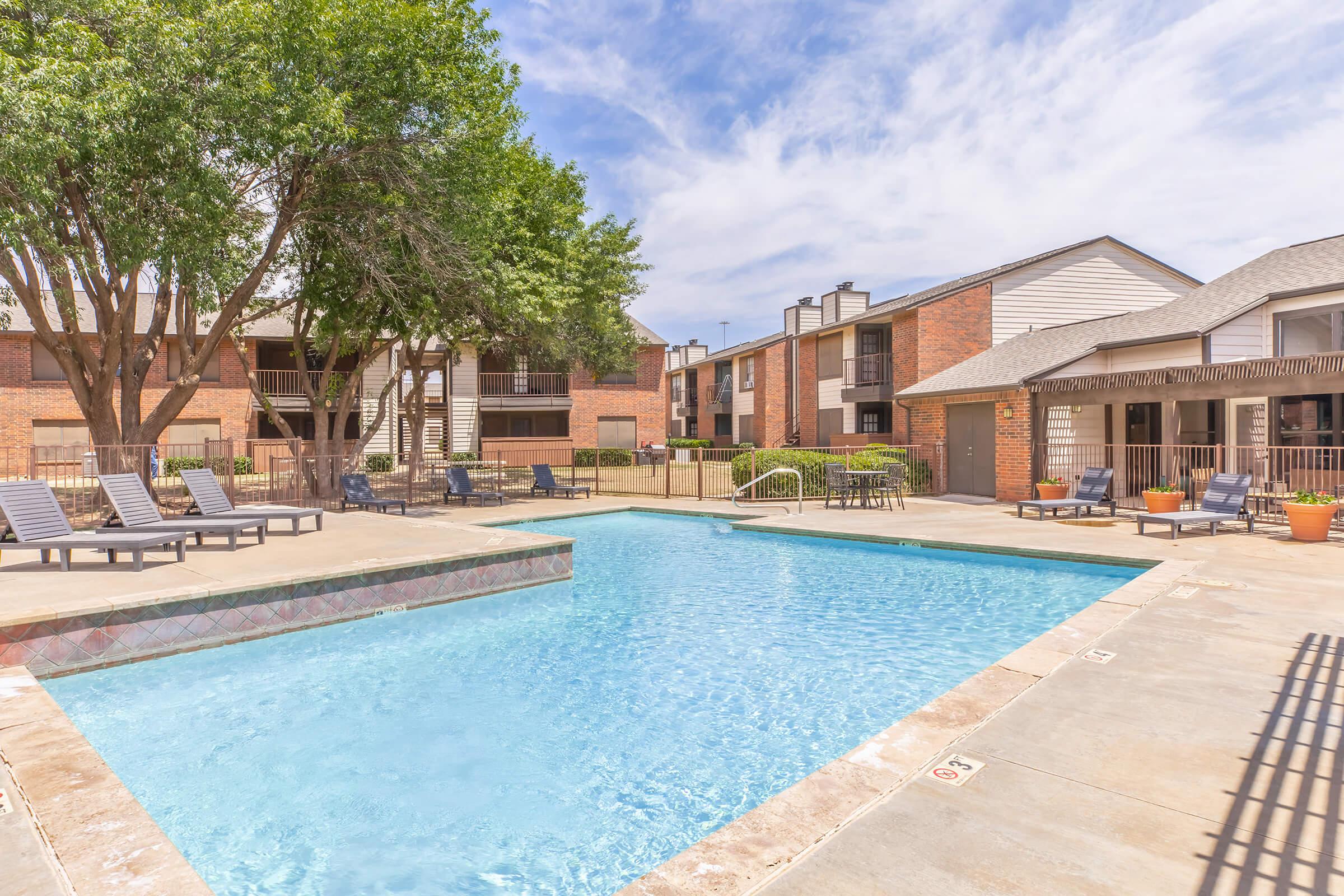 A bright outdoor swimming pool surrounded by lounge chairs, with lush green trees nearby. The pool area is bordered by brick apartment buildings under a blue sky with fluffy clouds, creating a welcoming and relaxing atmosphere.