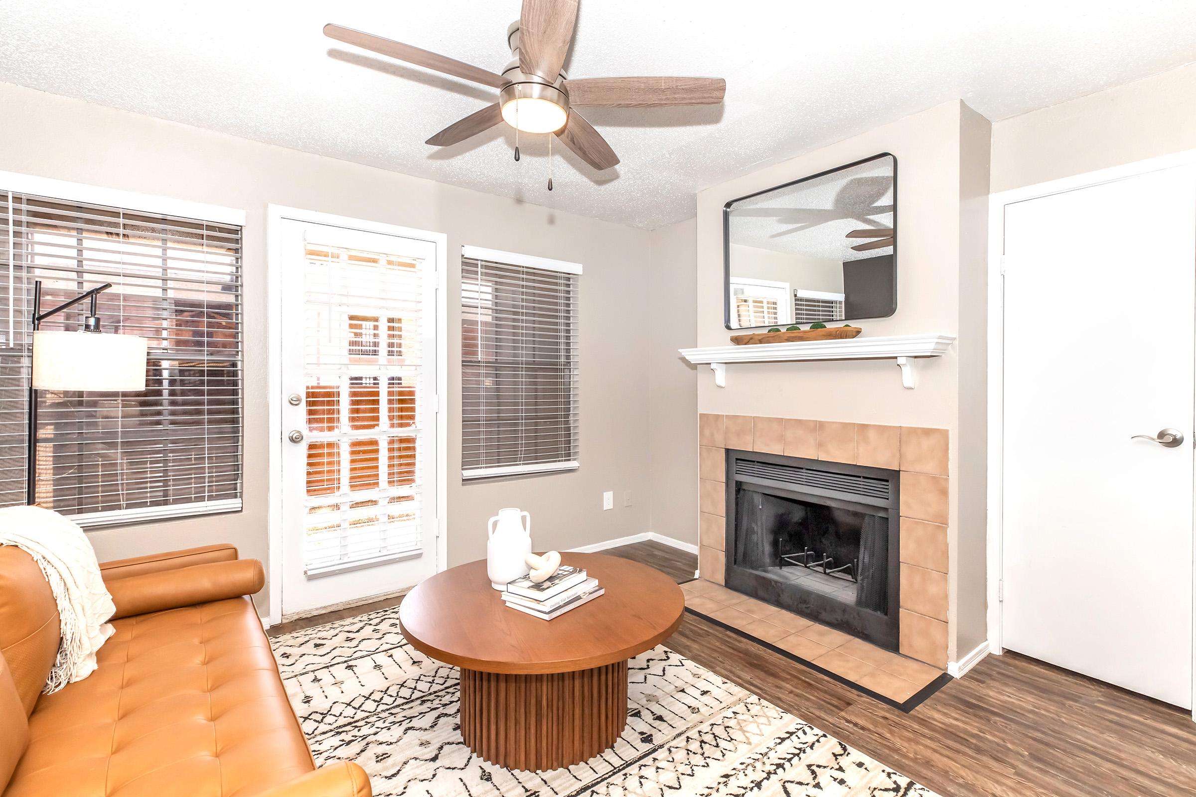 A cozy living room featuring a brown leather couch, a round wooden coffee table, and a fireplace. There are large windows with blinds letting in natural light. A ceiling fan hangs above, and a mirror is mounted on the wall above the fireplace, creating a warm and inviting atmosphere.