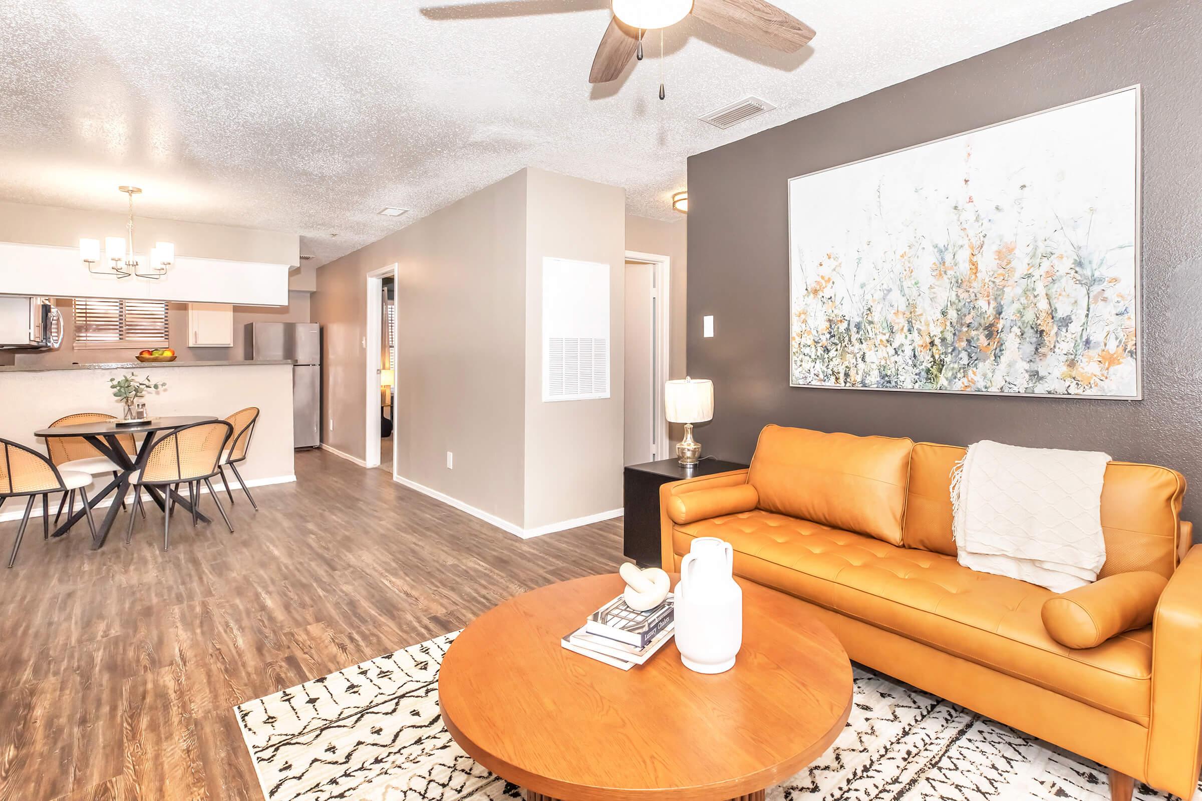 A modern living area featuring a stylish orange leather sofa, a round wooden coffee table, and decorative items. In the background, there's a dining area with a round table and chairs. The walls are painted in neutral tones, complemented by a large piece of artwork. Bright, natural light enhances the inviting atmosphere.