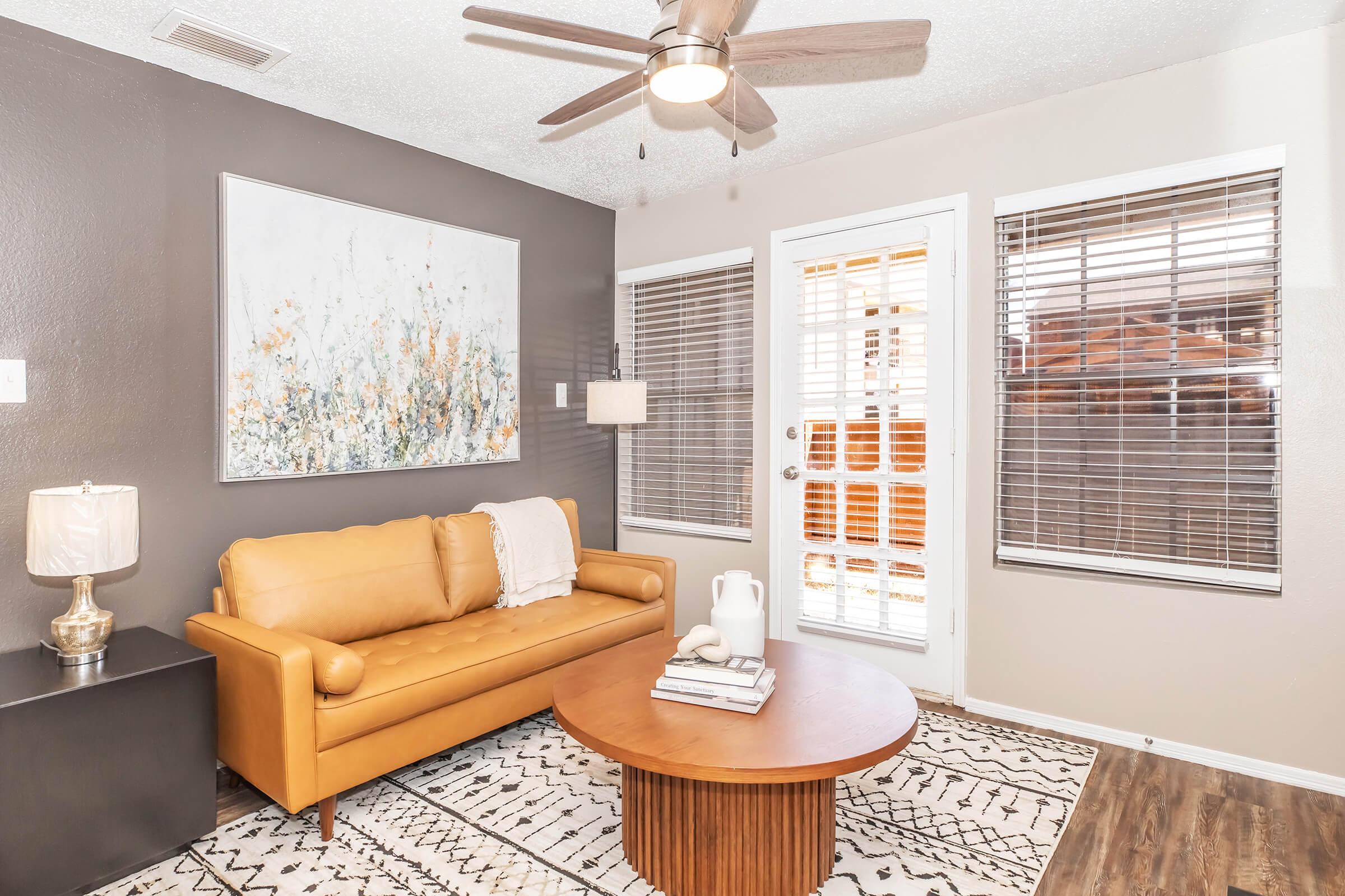 A cozy living room featuring a tan leather sofa, a round wooden coffee table with decorative items, and a patterned rug. The walls are painted in a dark shade, and there's a large artwork piece above the sofa. Double doors with blinds provide access to an outdoor space, and a ceiling fan hangs above.