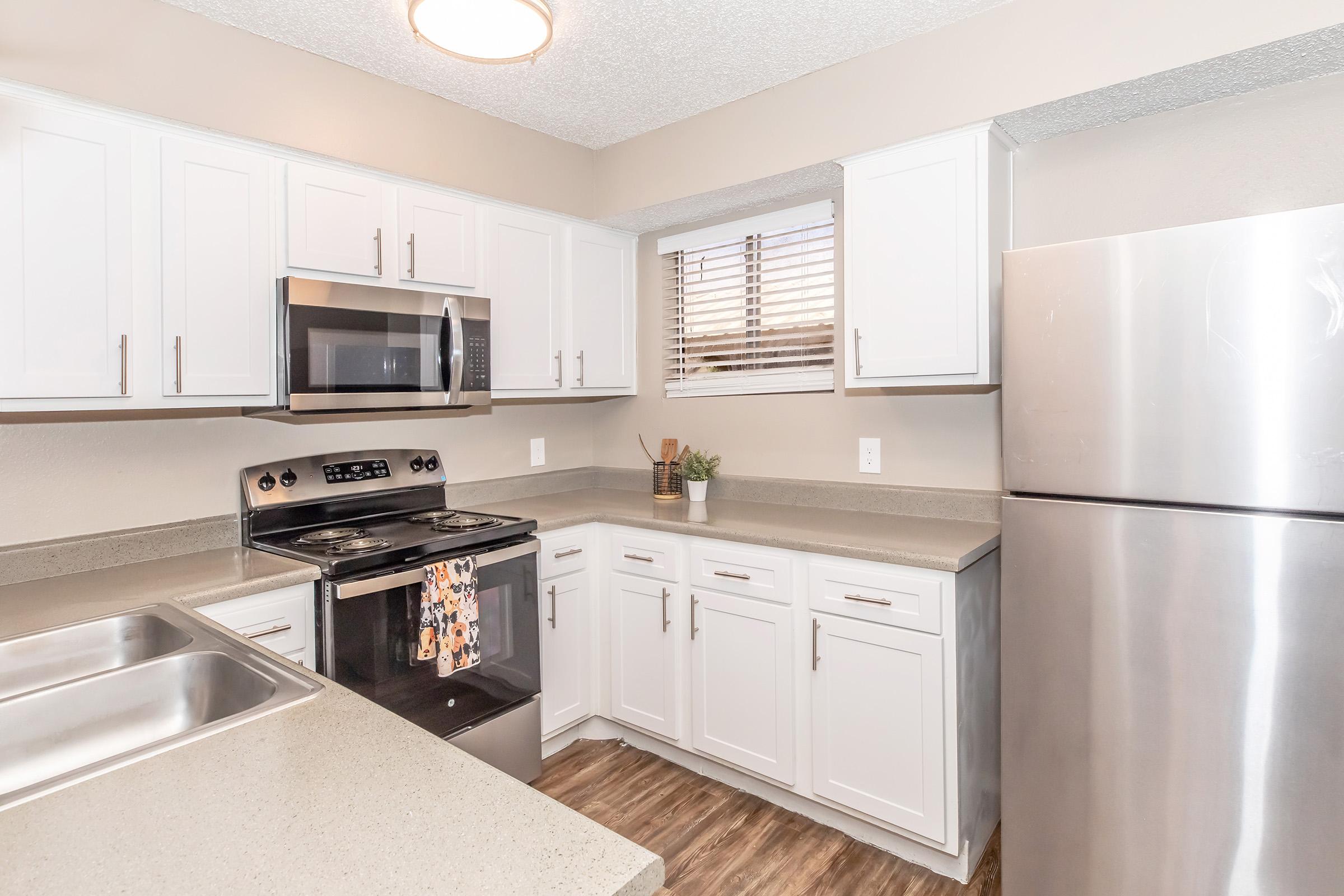 Modern kitchen featuring white cabinets, stainless steel appliances including a microwave and refrigerator, and a gray countertop. Natural light comes in through a window with blinds. There’s a sink and an organized layout, creating a bright and inviting space for cooking.