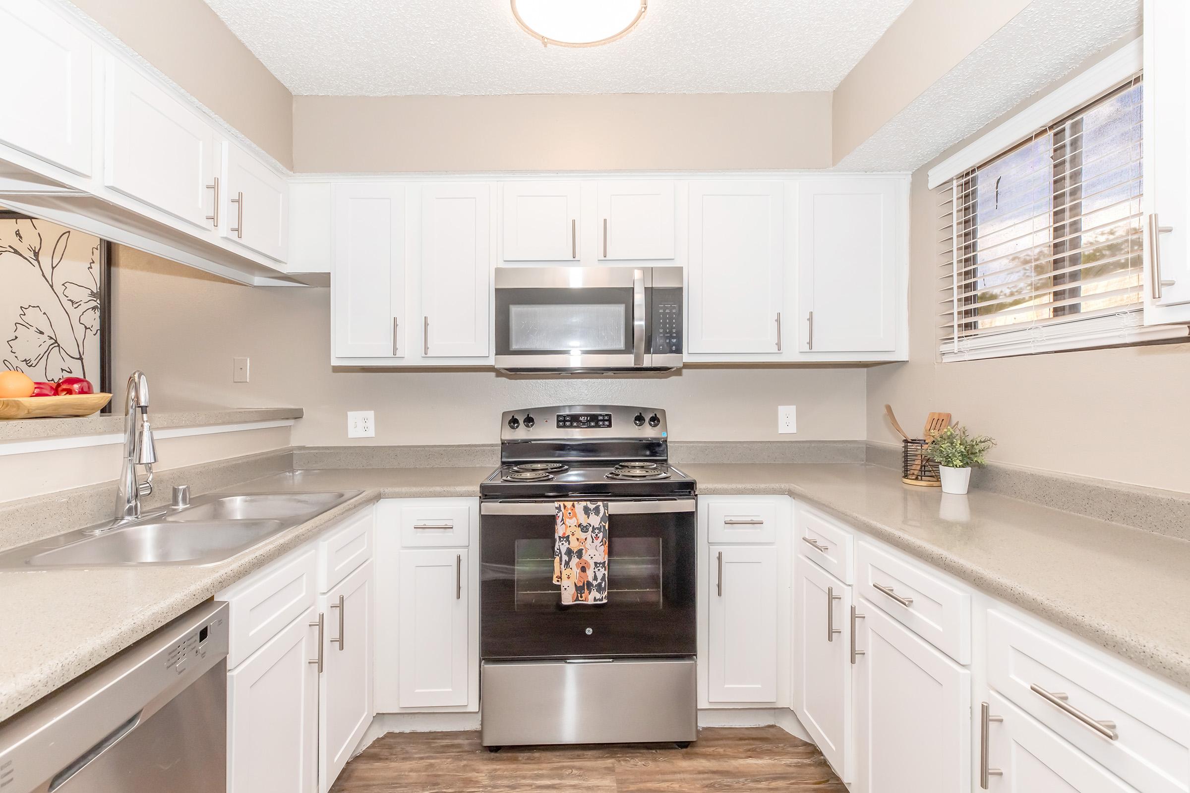 Bright and modern kitchen featuring white cabinets, a stainless steel stove and microwave, double sink, and a window with blinds. A bowl of apples is on the counter, adding a pop of color. The countertop has a light gray finish, and the floor is wood-like, creating a warm and inviting atmosphere.