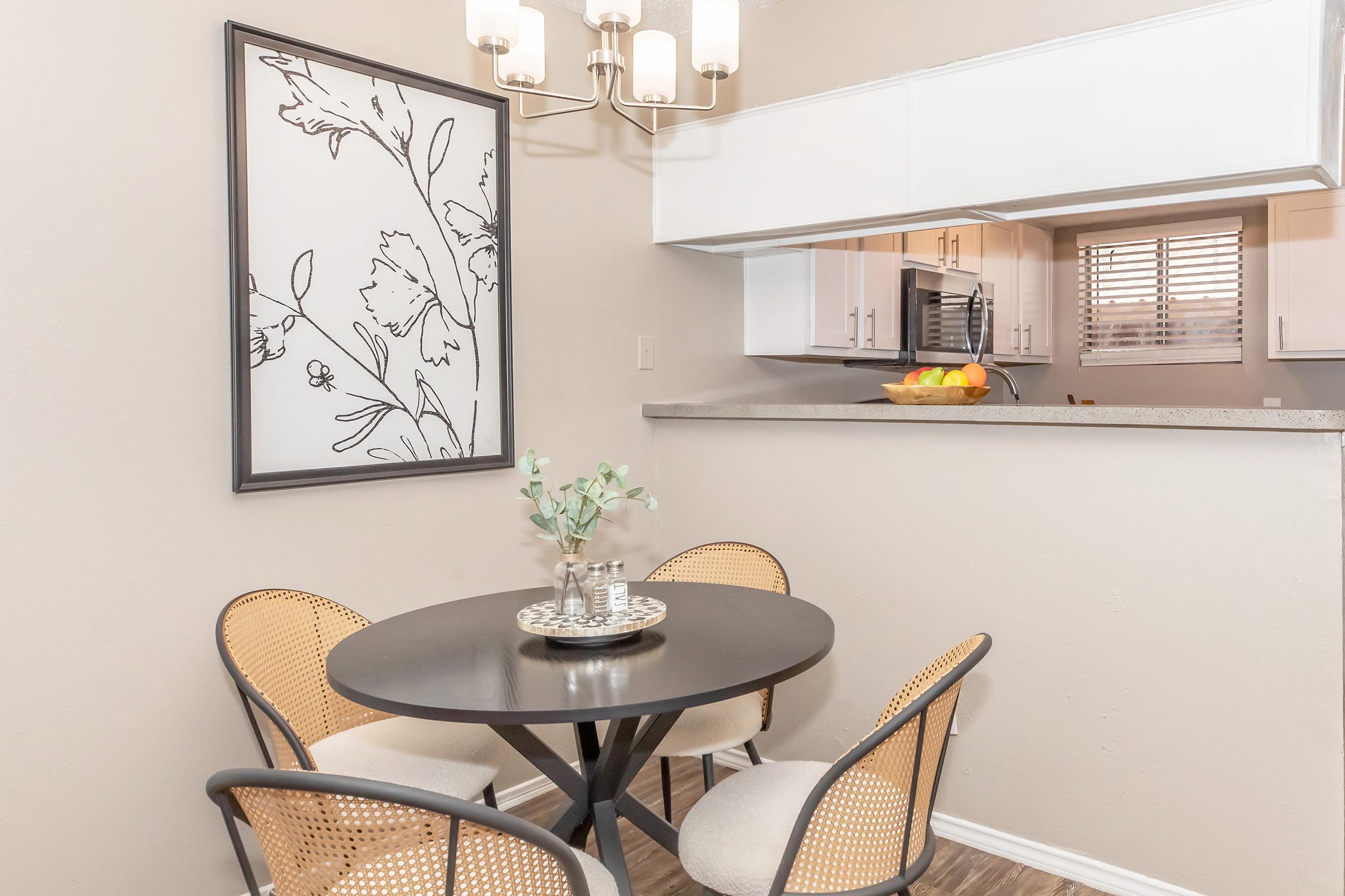 A cozy dining area featuring a round black table with four chairs and a decorative vase. A framed botanical print hangs on the wall, and a light fixture above adds warmth. The adjacent kitchen is visible, showcasing white cabinets and a bowl of fruit on the counter. Natural light filters through window blinds.