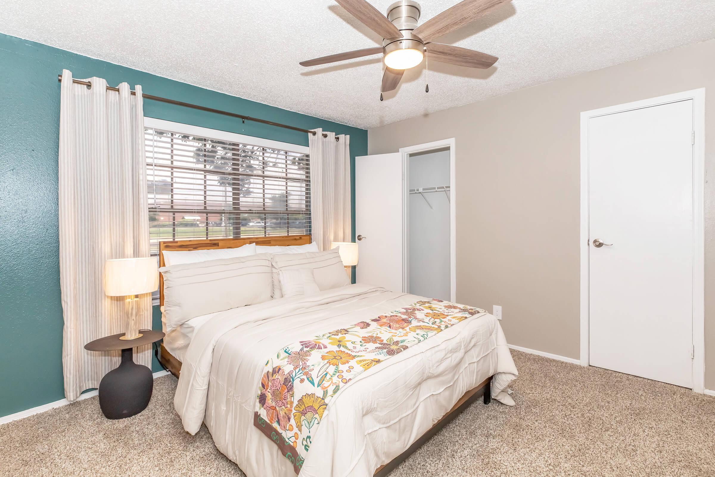A cozy bedroom featuring a queen bed with a floral patterned cover, two bedside lamps, and a ceiling fan. The walls are painted in a soothing teal and light beige. Natural light streams through a window with semi-sheer curtains, and a closet door is visible in the background. The carpeted floor adds warmth to the space.