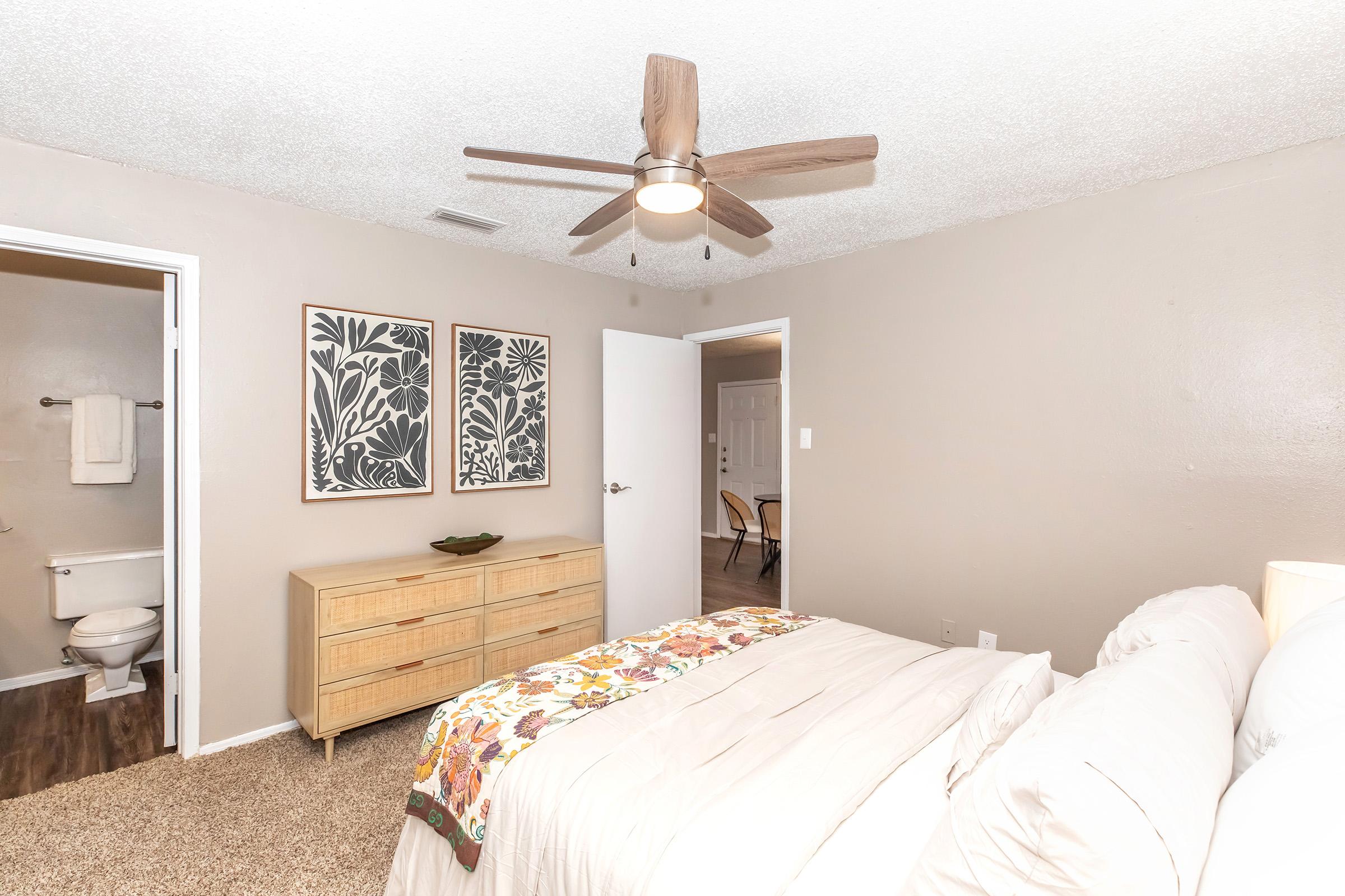 A cozy bedroom featuring a bed with a light-colored blanket, wooden dresser, and framed botanical wall art. A ceiling fan with wooden blades hangs above. There are two doors; one leads to a bathroom and the other to the rest of the home. The walls are painted in a soft beige hue, creating a warm, inviting atmosphere.