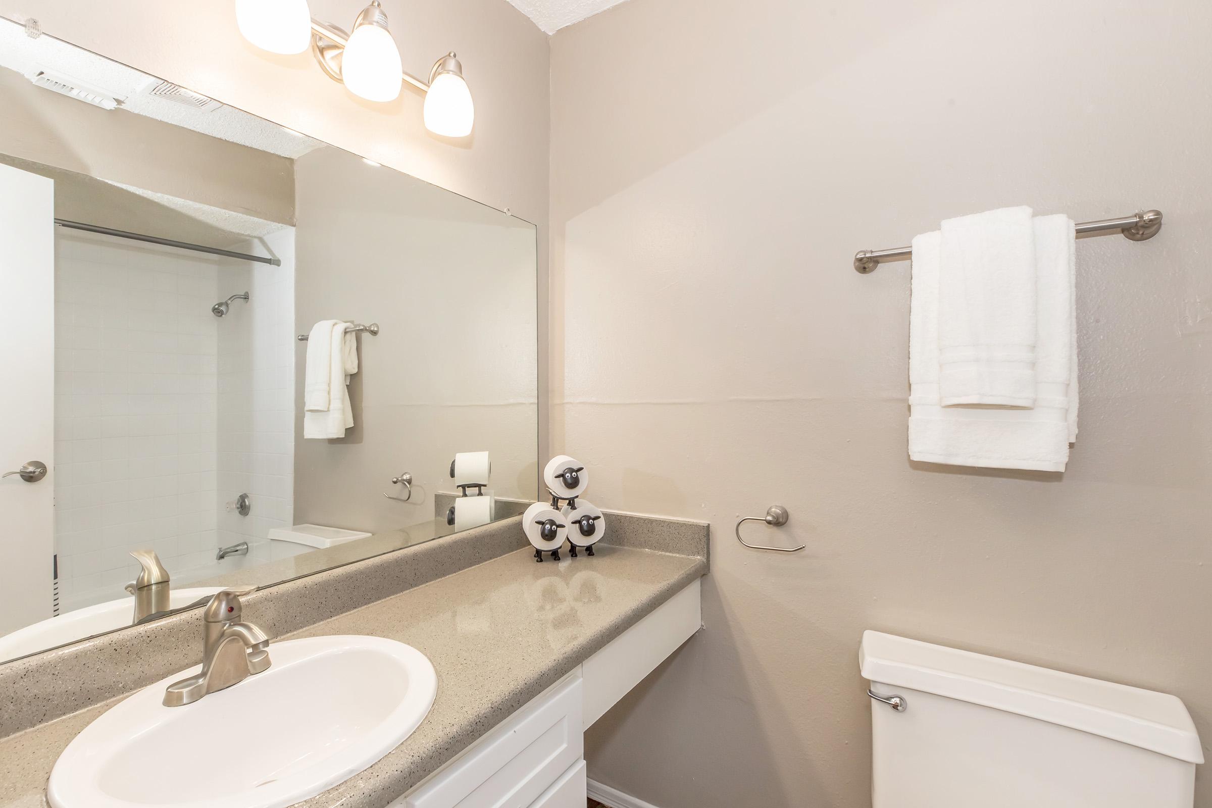A clean bathroom featuring a sink with a modern faucet, a large mirror above the sink, and a white countertop. Two neatly folded white towels hang on a towel bar. A shower curtain is visible in the background, and a compact toilet is positioned next to the sink area, creating a tidy and organized space.