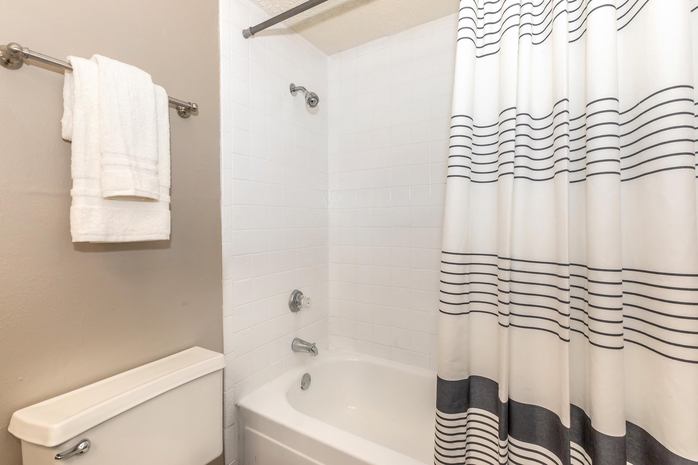 A clean bathroom featuring a white bathtub with a showerhead, a white shower curtain with black stripes, and a towel rack with neatly hung white towels. The walls are painted a light color, and there's a toilet adjacent to the tub.