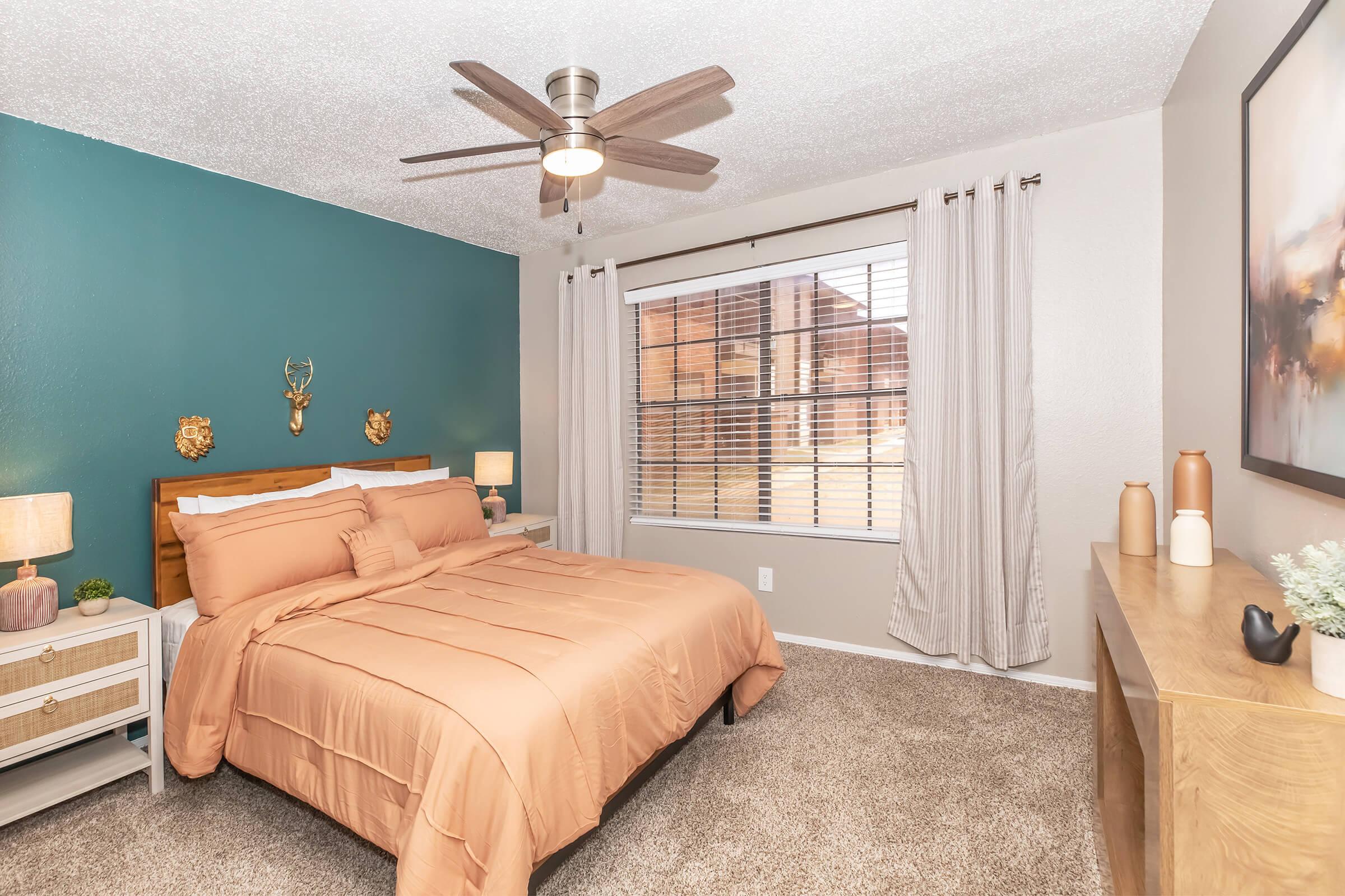 Cozy bedroom featuring a queen-sized bed with light brown bedding, a teal accent wall, and two decorative wall mounts. The room includes bedside tables with lamps, a window with sheer curtains, and a wooden dresser against one wall. A ceiling fan adds to the comfort of the space.