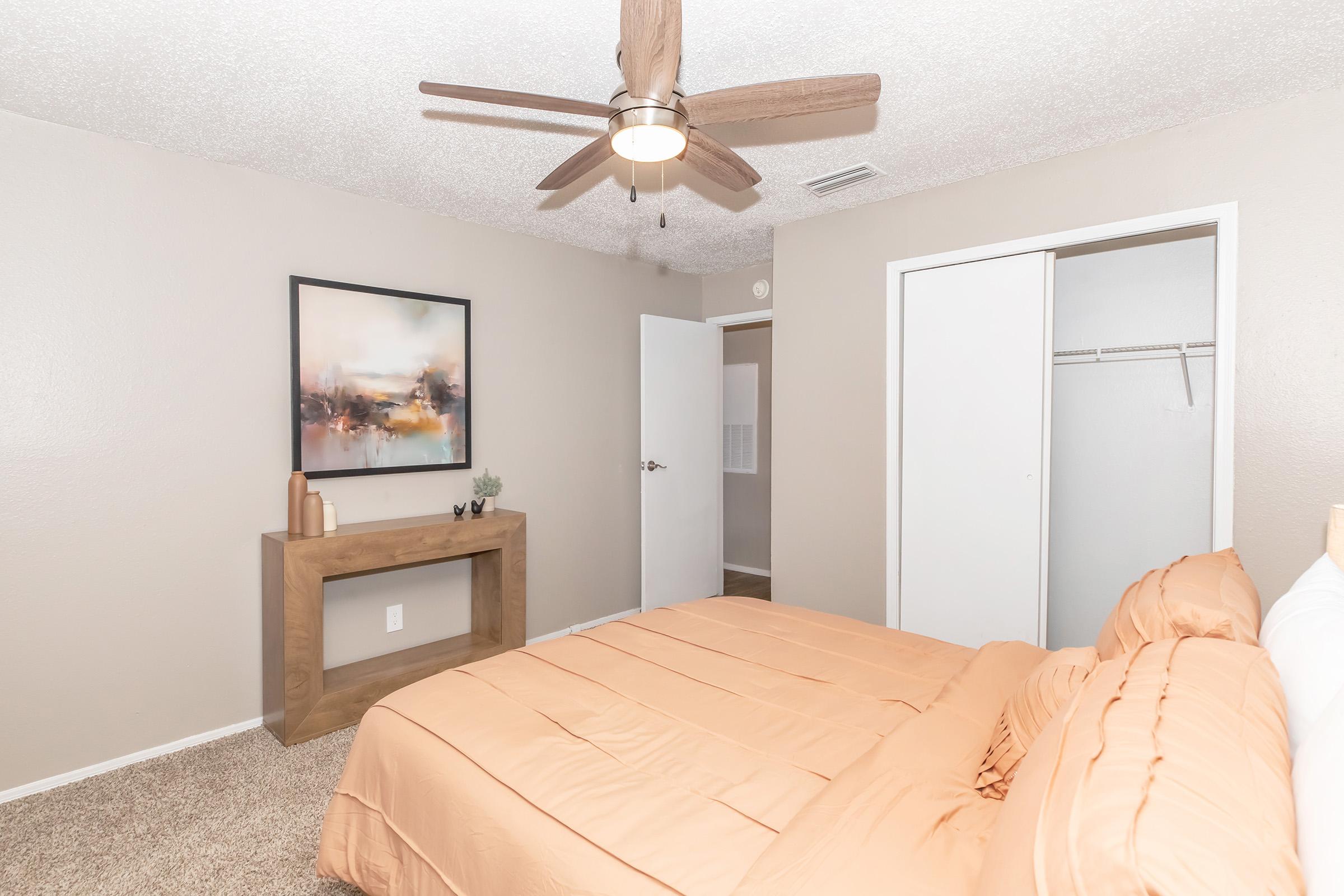 A cozy bedroom featuring a queen-sized bed with an orange bedspread, a modern wooden console table, a ceiling fan, and a piece of abstract wall art. The room has light-colored walls and carpet, with a closet visible in the background and a door leading to another space.
