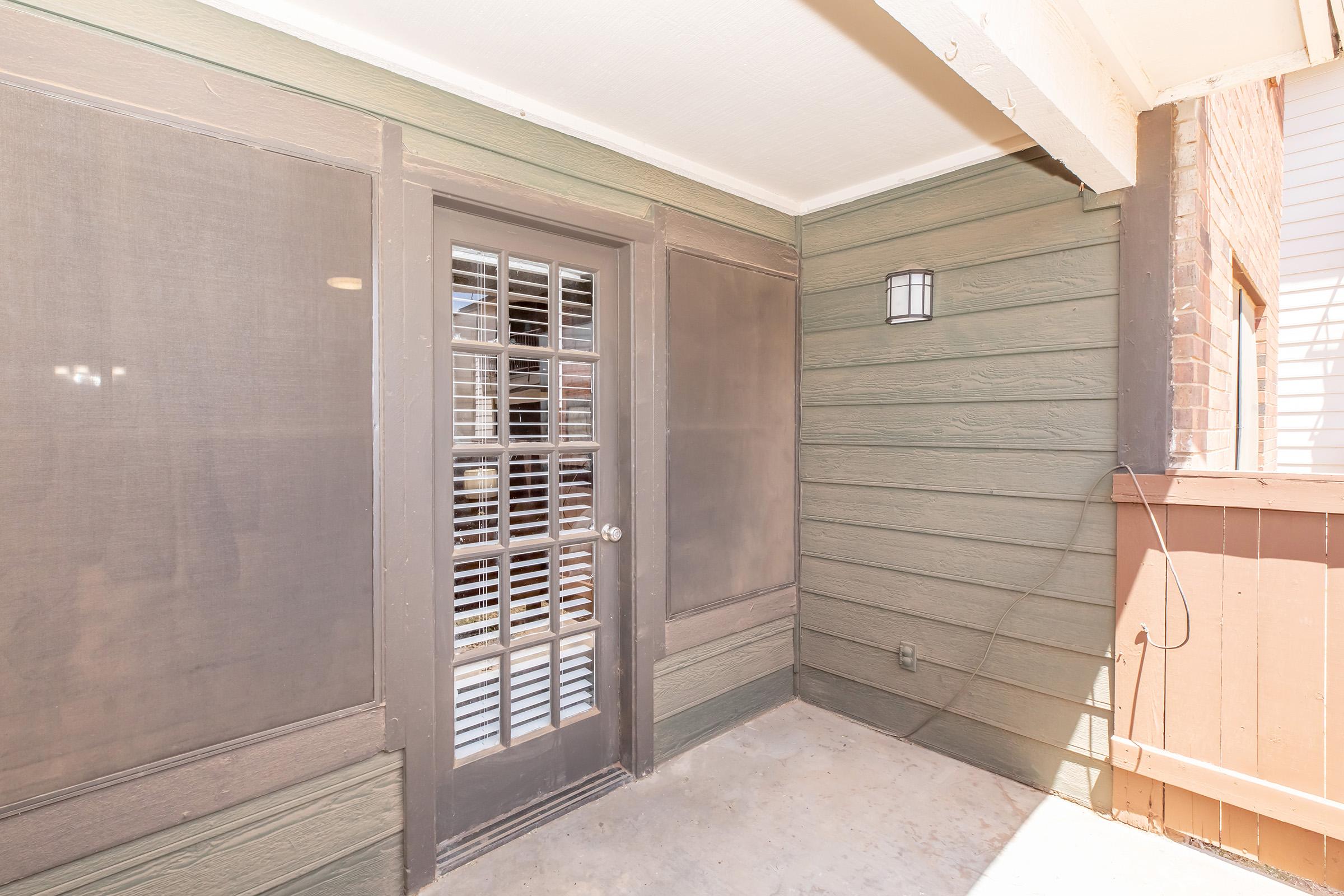 A view of a covered outdoor patio area with a door featuring large glass panes and a decorative light fixture on the wall. The walls are made of a greenish-brown wood material, and the floor is concrete. The space is enclosed by screening, providing a shaded environment.