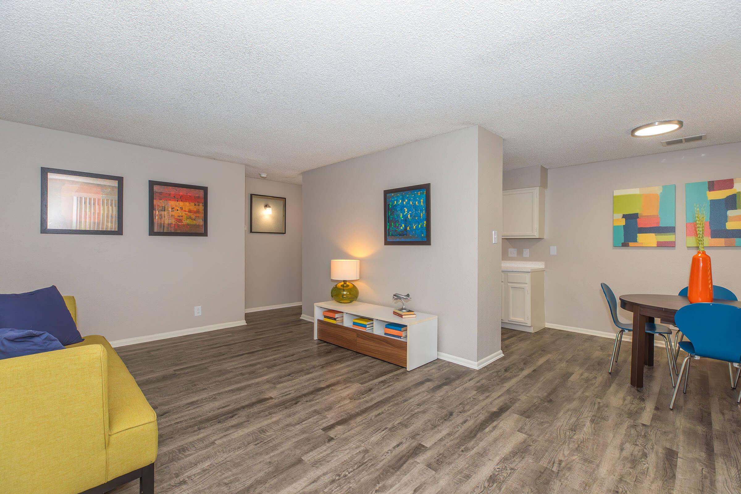 A modern living space featuring a cozy yellow sofa, a small coffee table with decorative items, and artwork on the walls. The room has a dining area with a round table and colorful chairs, set against a neutral-colored wall and wood-like flooring. Bright lighting enhances the inviting atmosphere.