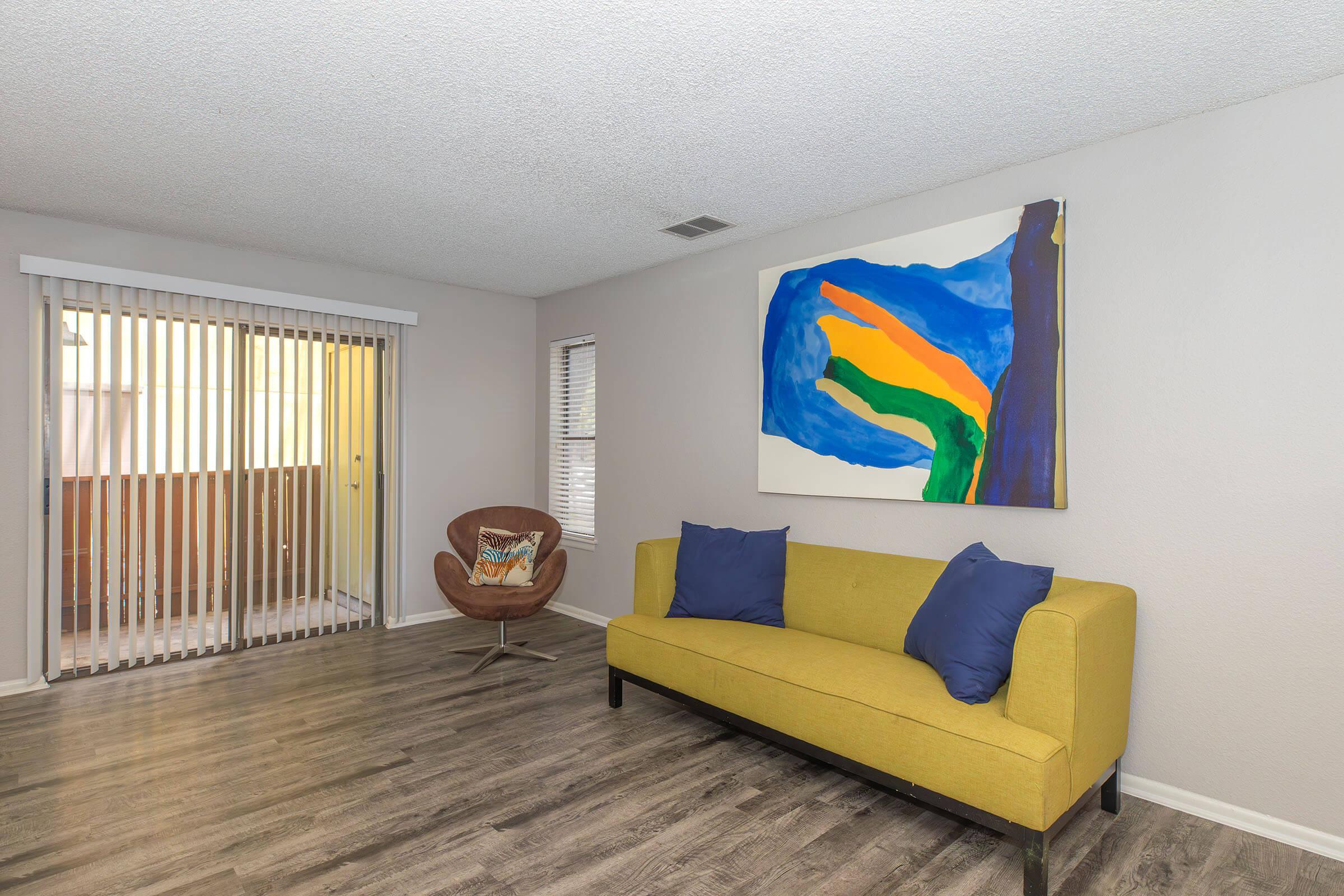 A minimalist living room featuring a green sofa with blue pillows, an abstract painting with bold colors hanging on the wall, and a brown chair. There are large sliding glass doors with vertical blinds, allowing natural light into the space. The flooring is a light wood finish.